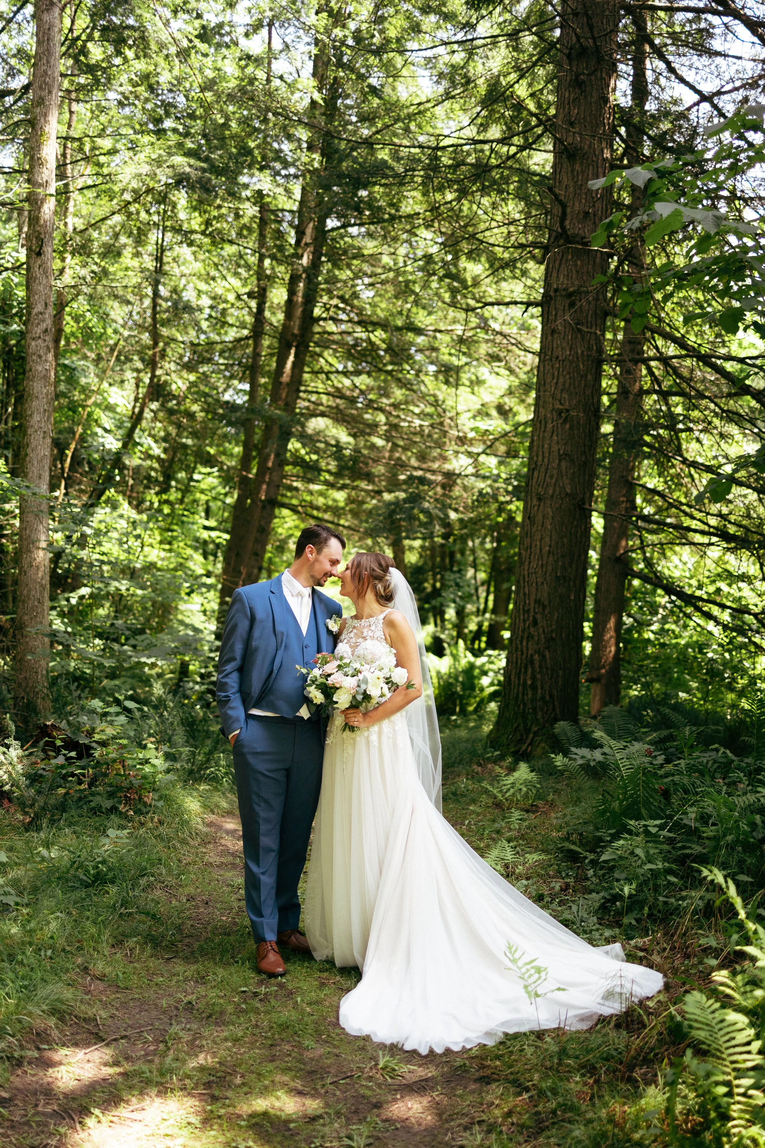 A bride and groom in wedding attire standing close together in a lush, sunlit forest, gazing at each other with noses touching, surrounded by tall trees and green foliage.