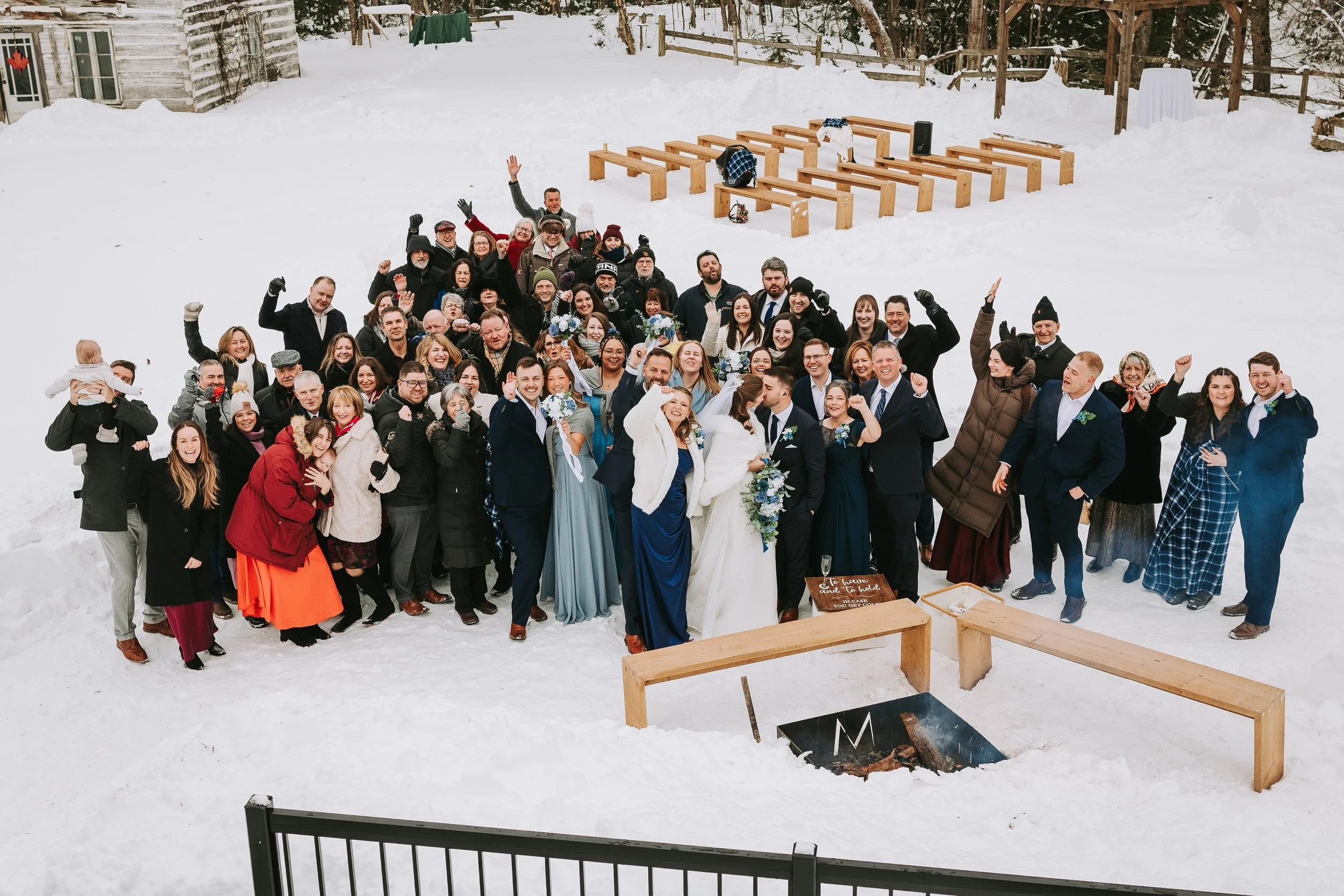 Group of people gathered outdoors in snow at a wedding, with some in formal attire, celebrating and smiling in front of a rustic wooden structure with benches and snow.