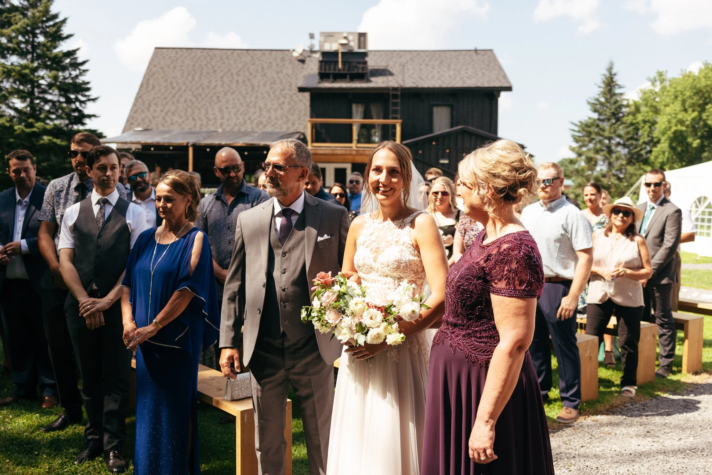 A bride holding a bouquet standing with her parents during a wedding ceremony outdoors on a sunny day, with guests in the background and a house behind them.