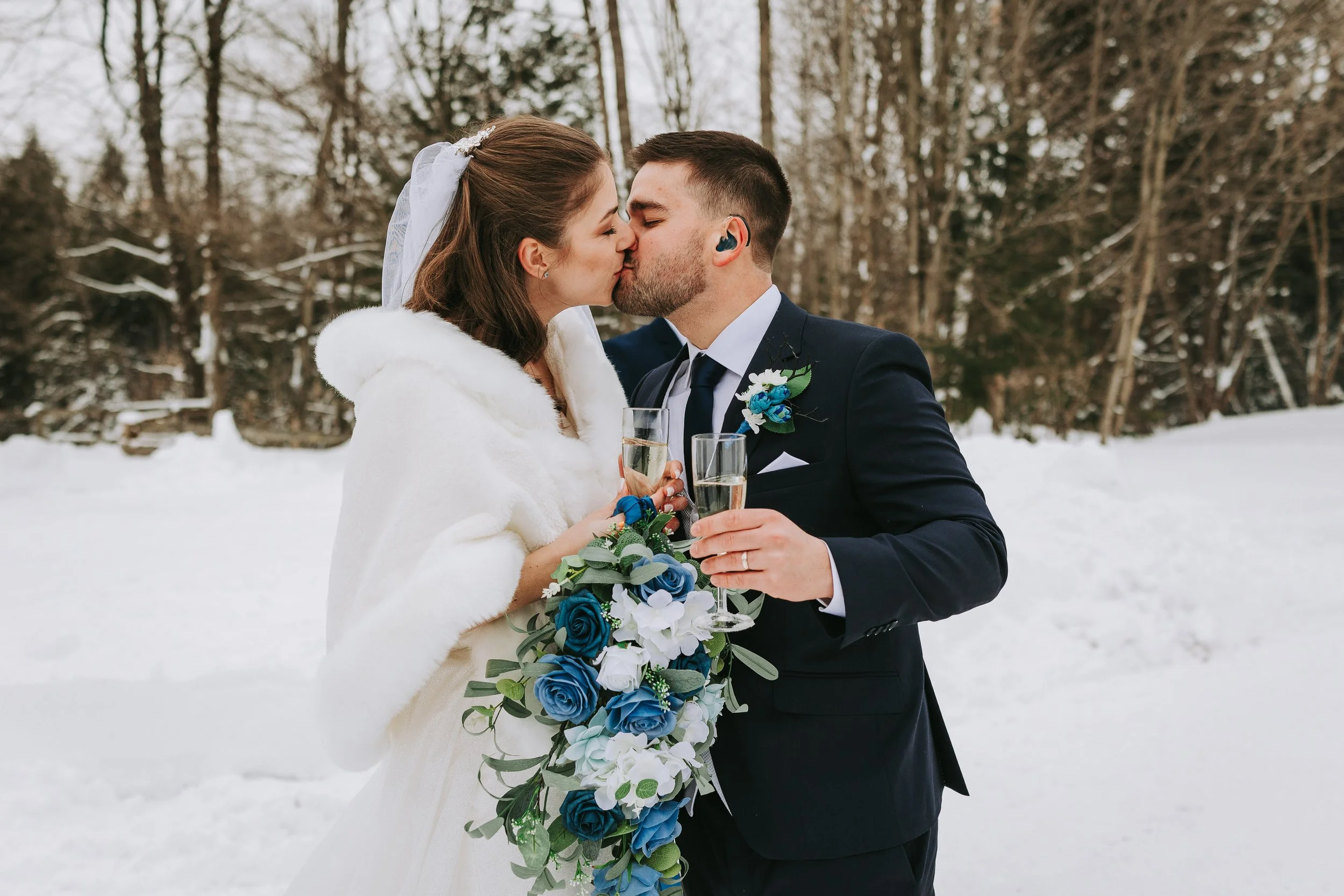 A bride and groom kiss while holding champagne flutes outdoors in a snowy winter landscape, with the bride wearing a white dress and fur cape, and the groom in a dark suit with a boutonnière.