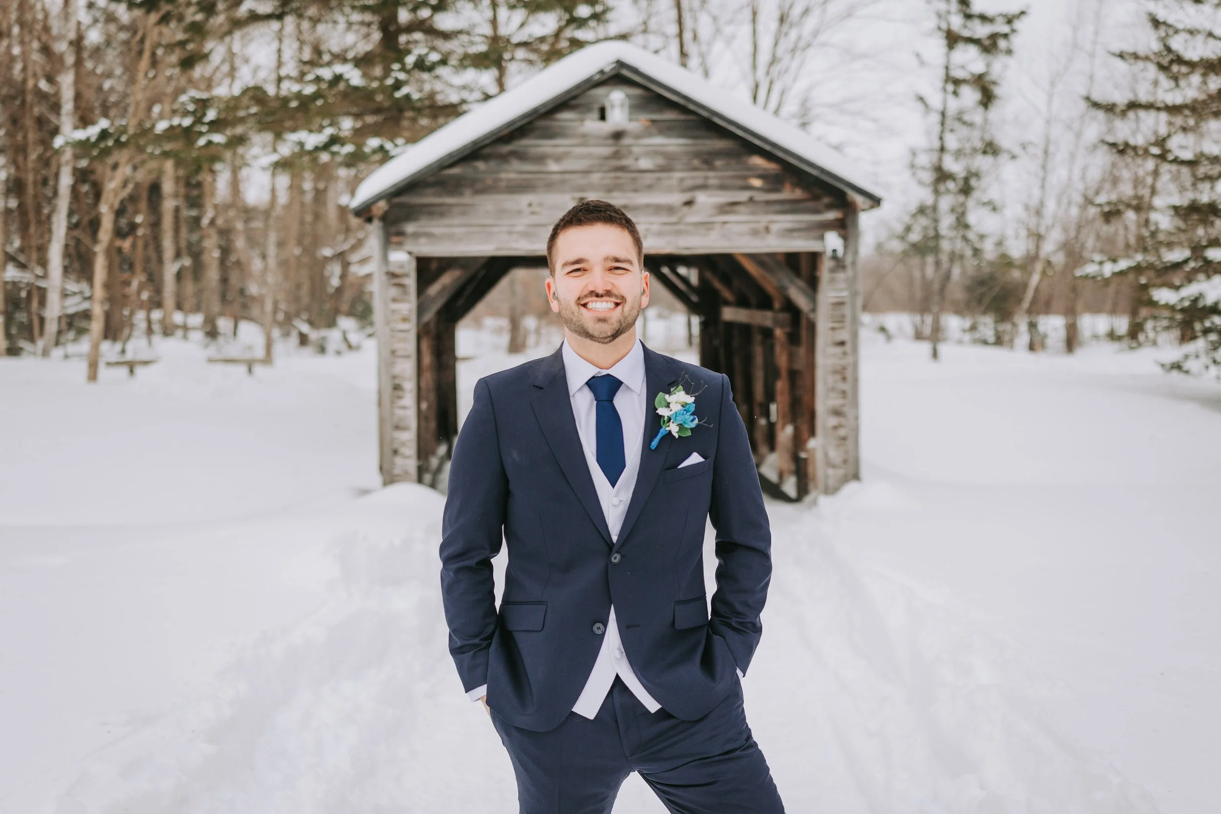 A man in a navy suit with a white shirt and blue tie standing outside in snow, smiling, with a rustic wooden cabin and snow-covered trees in the background.