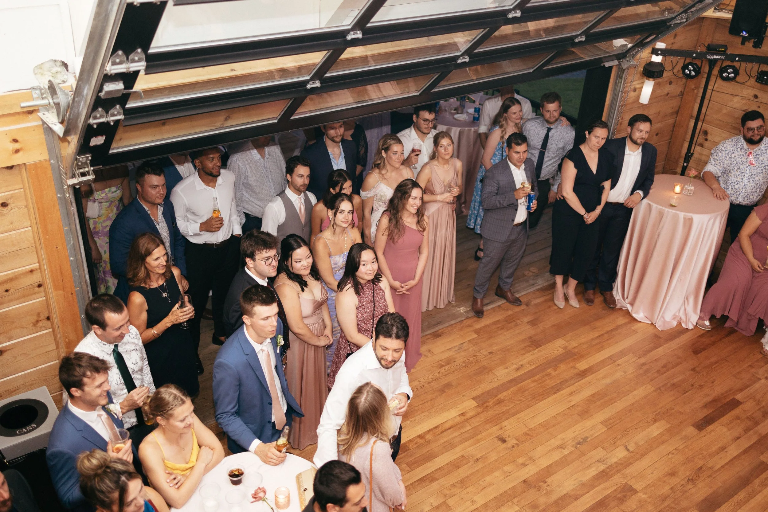 Group of people at a wedding reception in a ottawa wedding venue, some standing and some sitting at tables, with wooden floors and walls, some holding drinks.