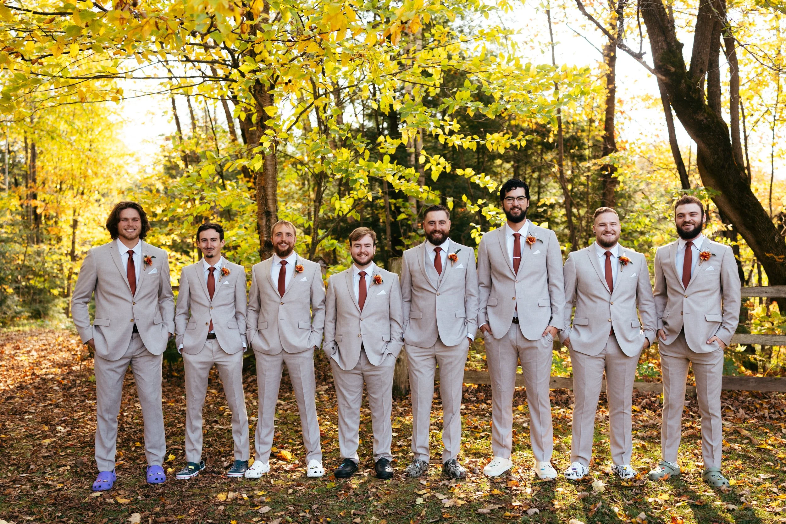 A group of nine men dressed in matching beige suits with white shirts and rust-colored ties standing outdoors in a wooded area with autumn leaves. They are smiling and standing side by side in a line, some with hands in pockets.