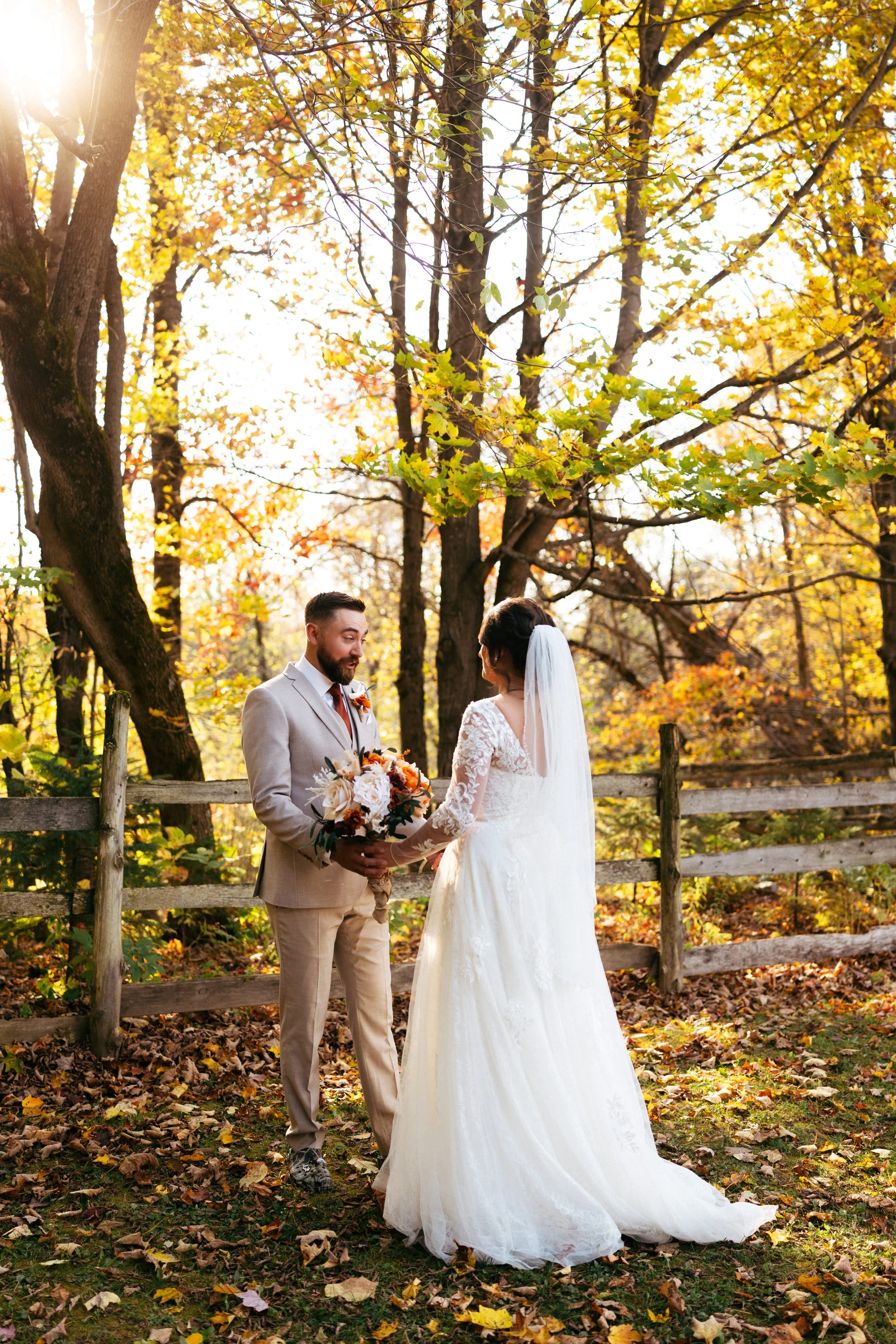 A bride and groom holding hands during an outdoor wedding in an autumn setting, surrounded by colorful fall leaves and trees.