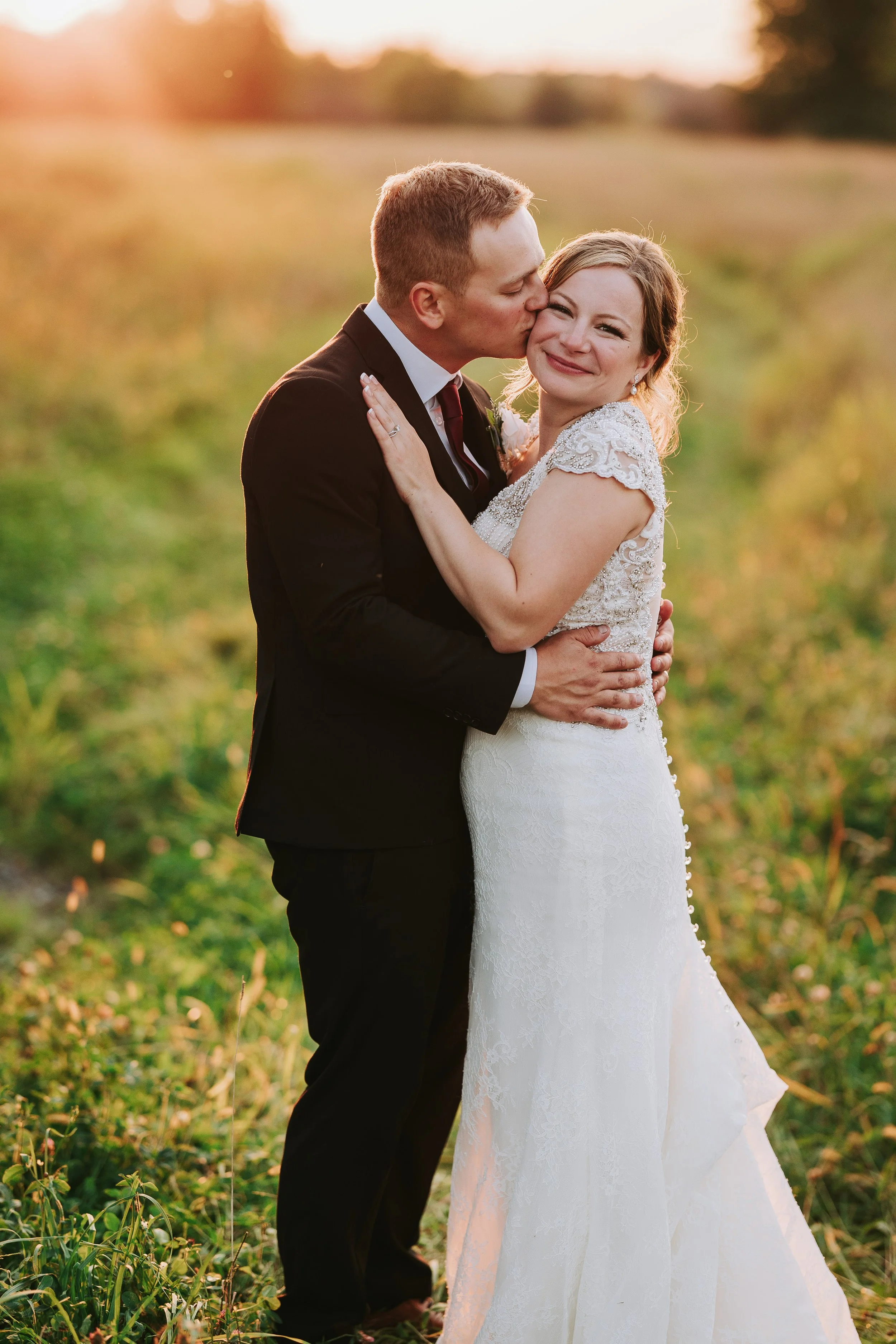 A newlywed couple embracing outdoors during sunset, with the groom kissing the bride on the cheek. The bride is wearing a lace wedding dress and smiling, while the groom is in a black suit and white shirt.