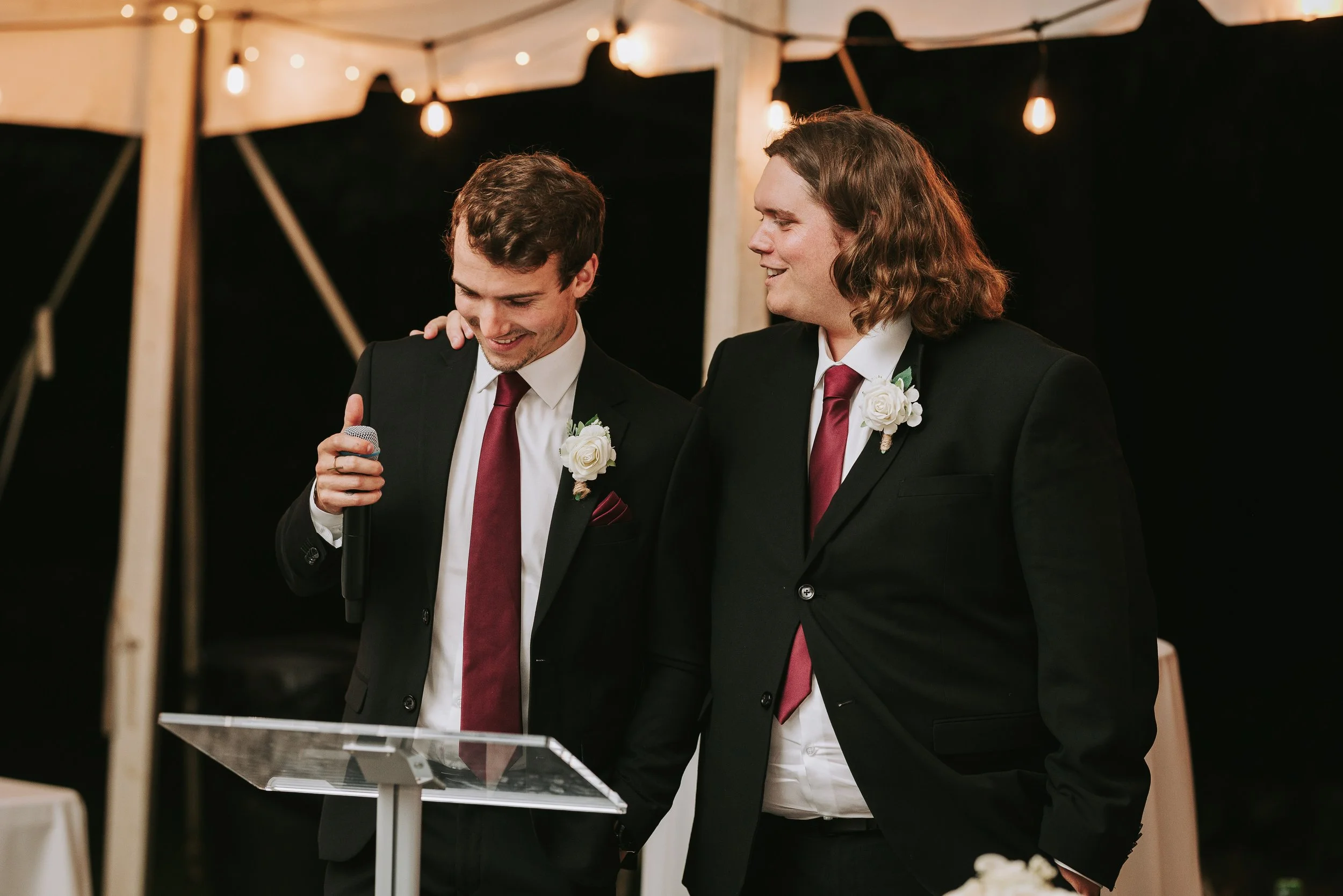 Two men in tuxedos, one with a microphone, standing close and smiling at a wedding reception under a tent with string lights.