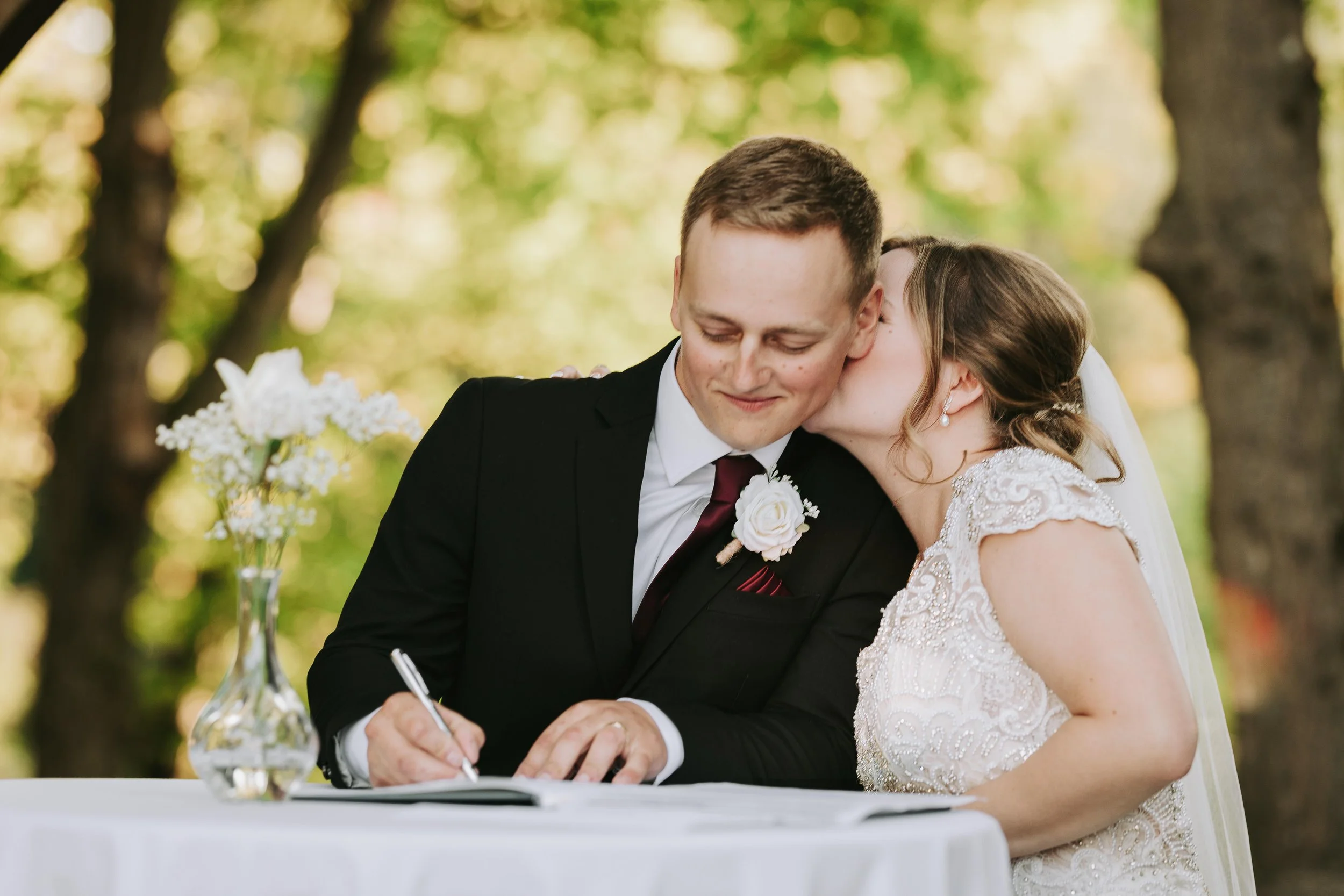 Bride kissing groom on the cheek while he signs a marriage document outdoors.