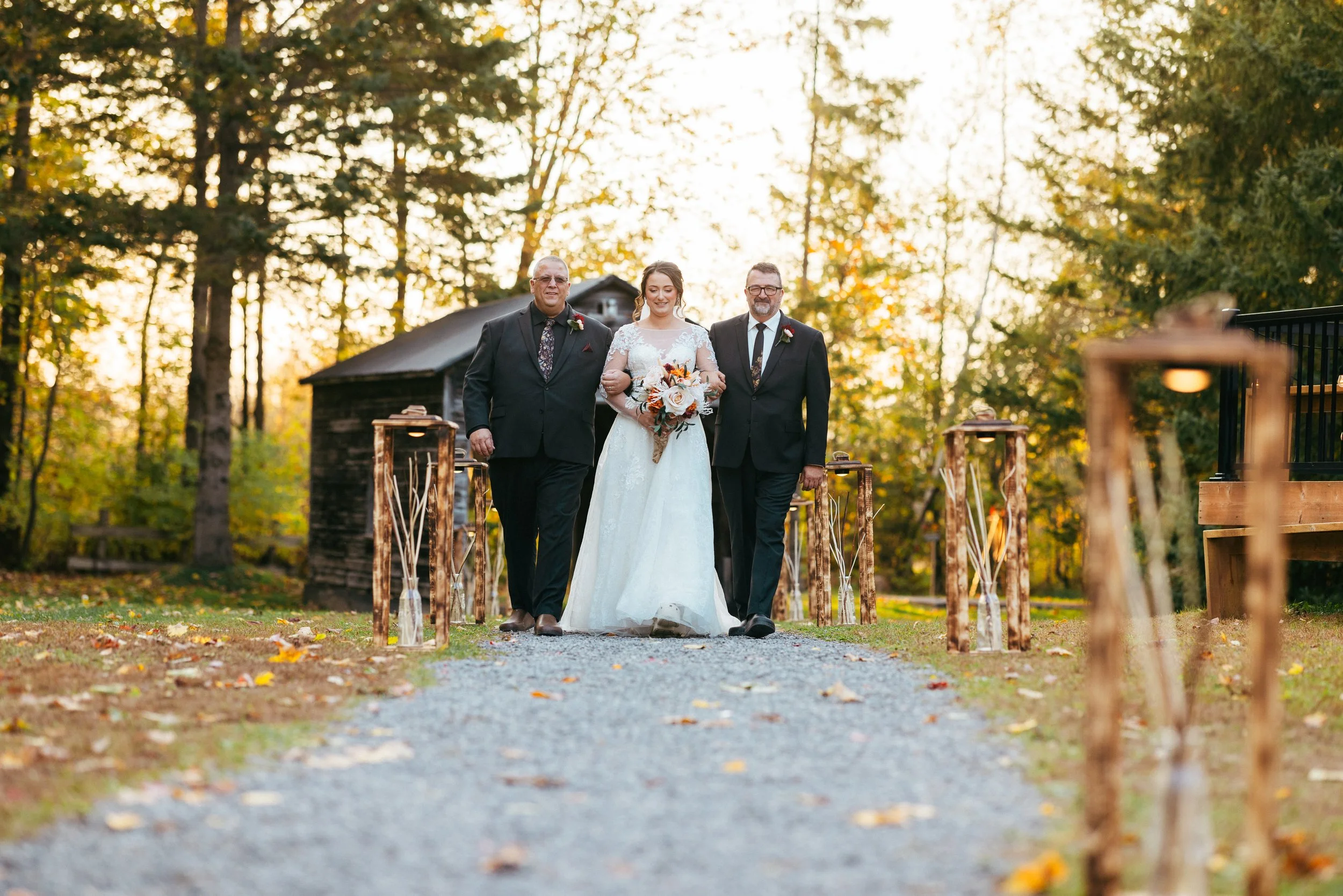 A bride walking down an outdoor aisle flanked by two men in suits, during a wedding ceremony in a wooded area at sunset.