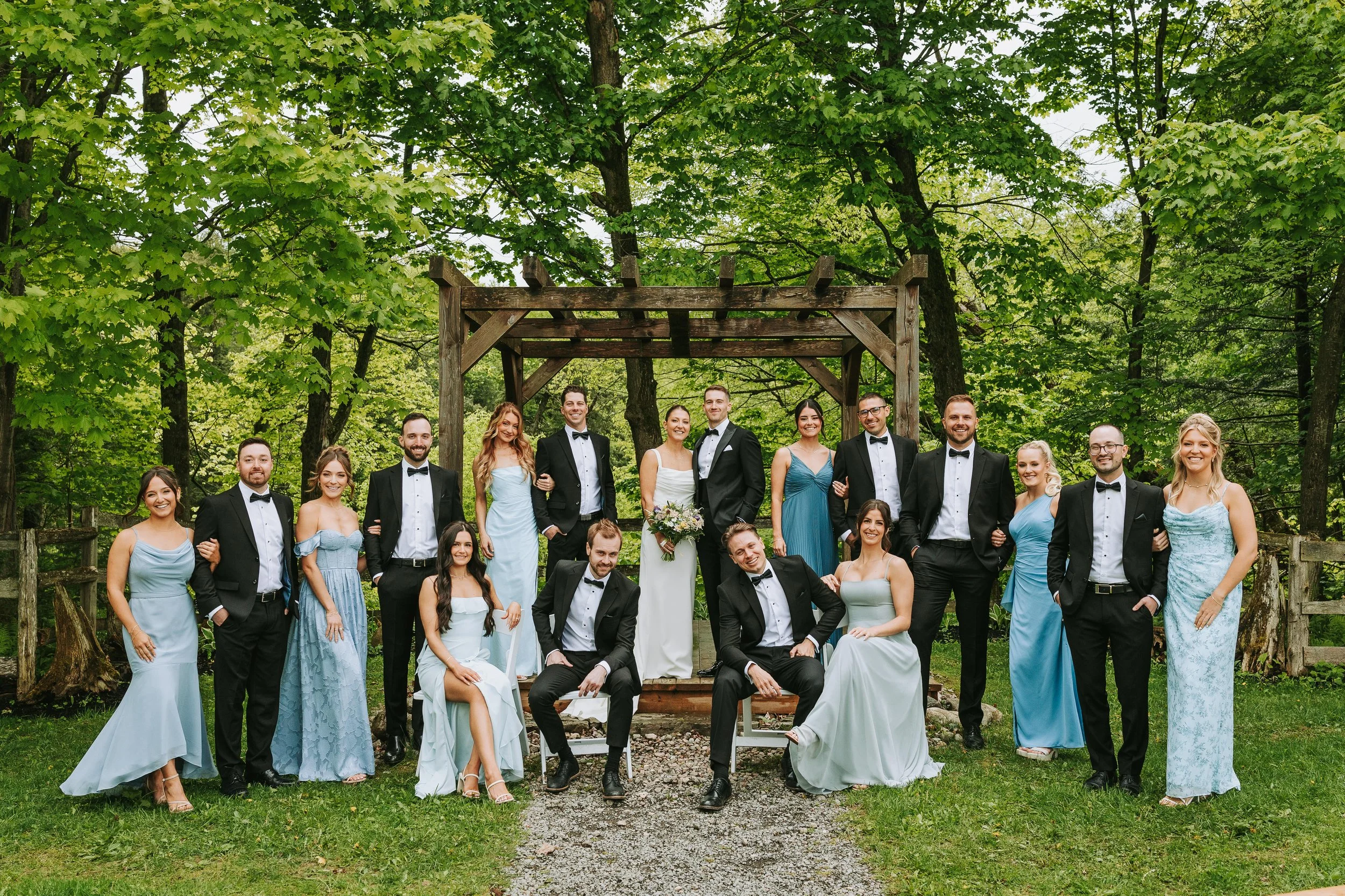 A group of people in formal attire posing outdoors in front of a wooden structure surrounded by trees.