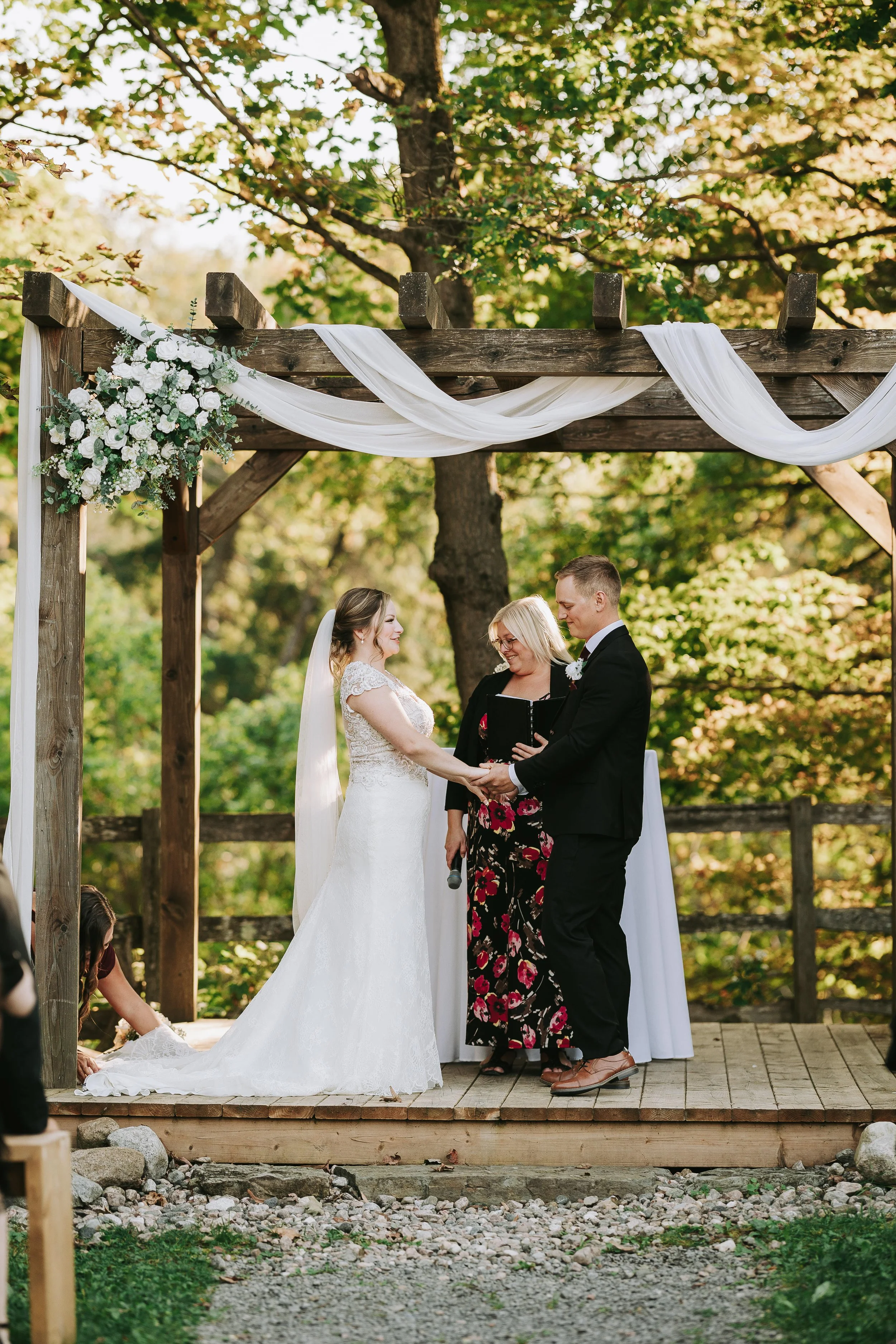 A couple gets married outdoors on a wooden platform under a wooden arch decorated with white flowers and fabric, with green trees in the background.