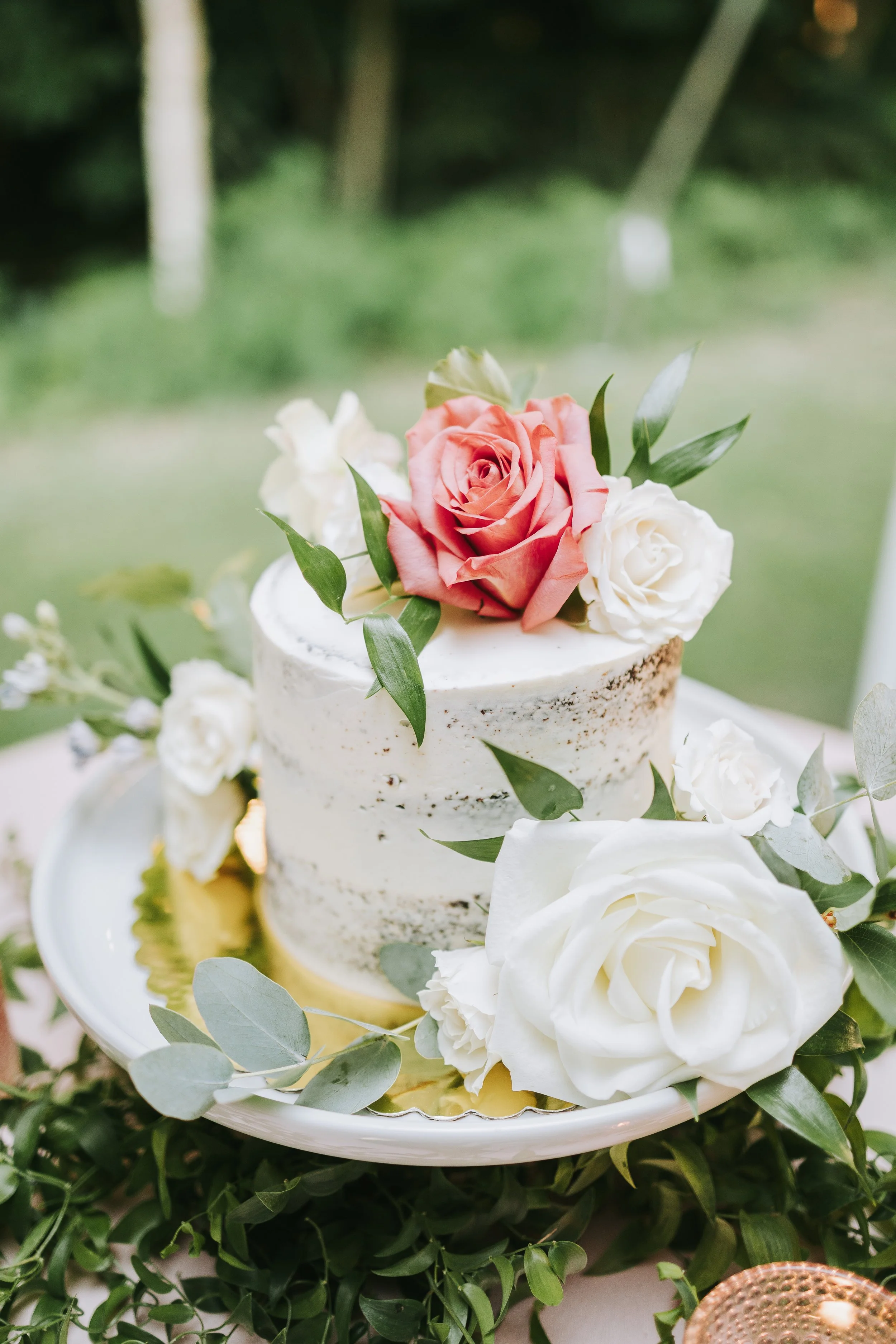 A semi-naked wedding cake decorated with pink and white roses and green leaves, placed on a white plate with greenery underneath, outdoors with a blurred green background.