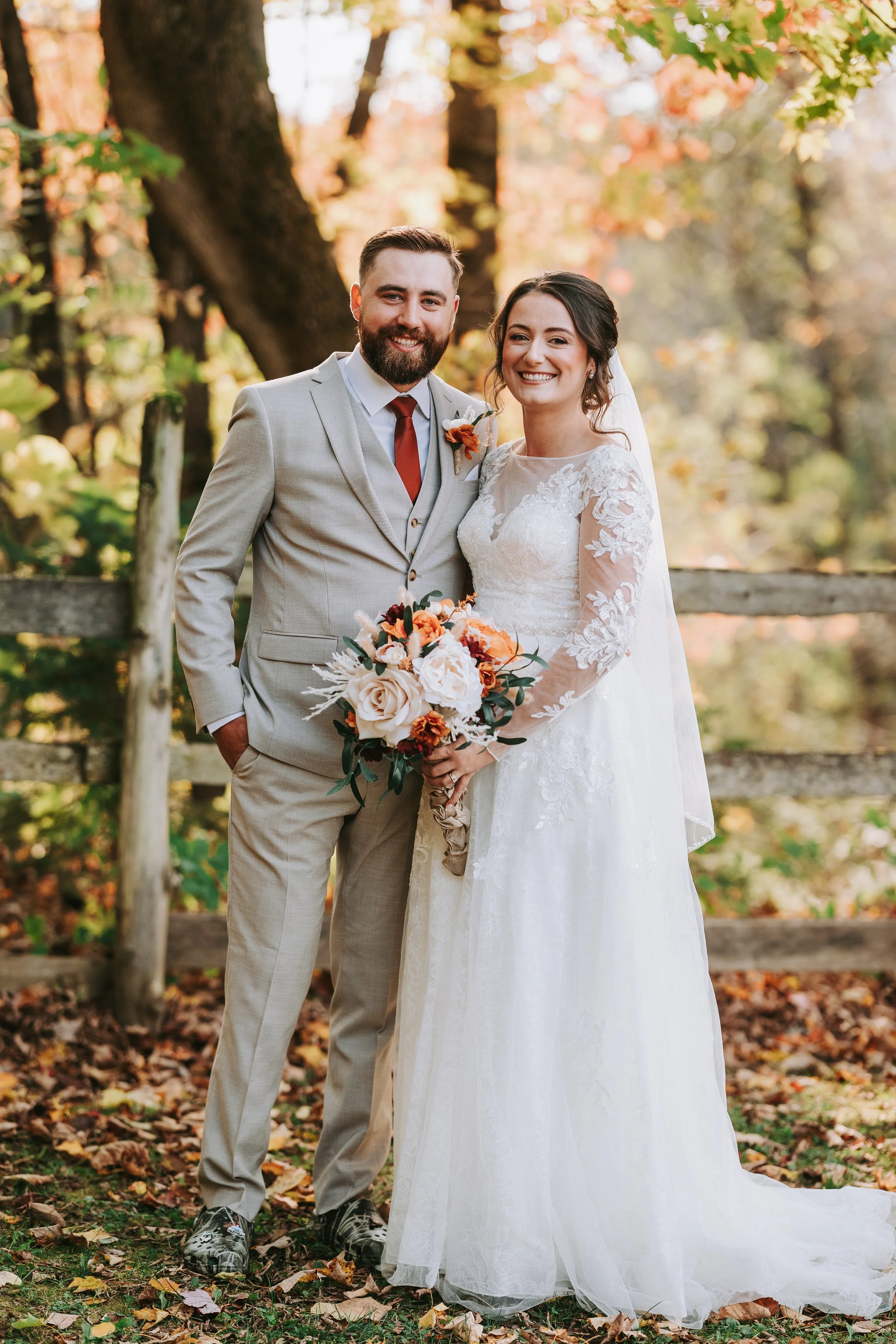 A newlywed couple standing outdoors in fall, smiling at the camera. The groom is wearing a light gray suit, a white shirt, and a red tie. The bride is in a white lace wedding dress holding a bouquet of peach, white, and dark red flowers. They are in 