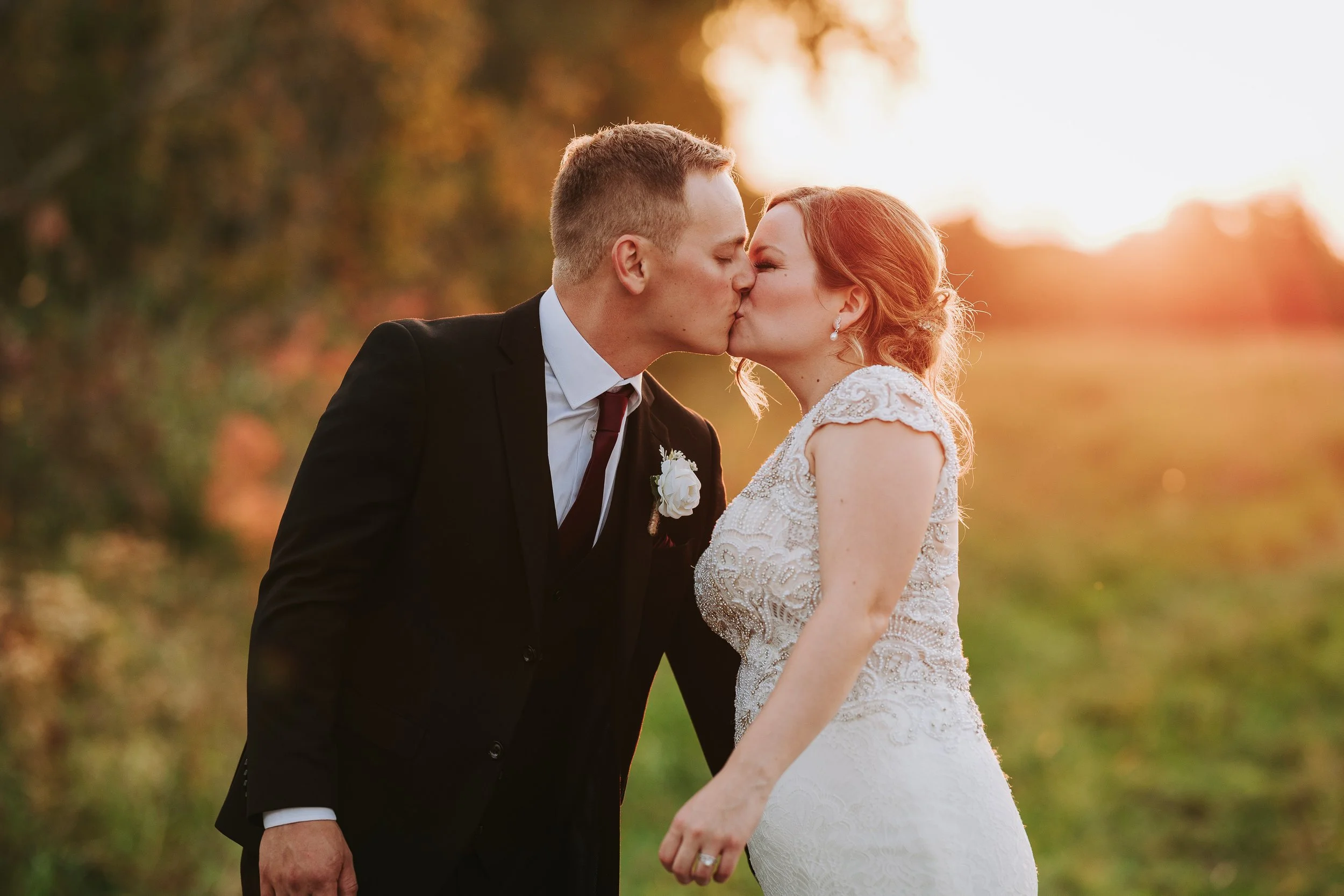A couple in wedding attire kissing outdoors at sunset, with trees and a glowing sky in the background.