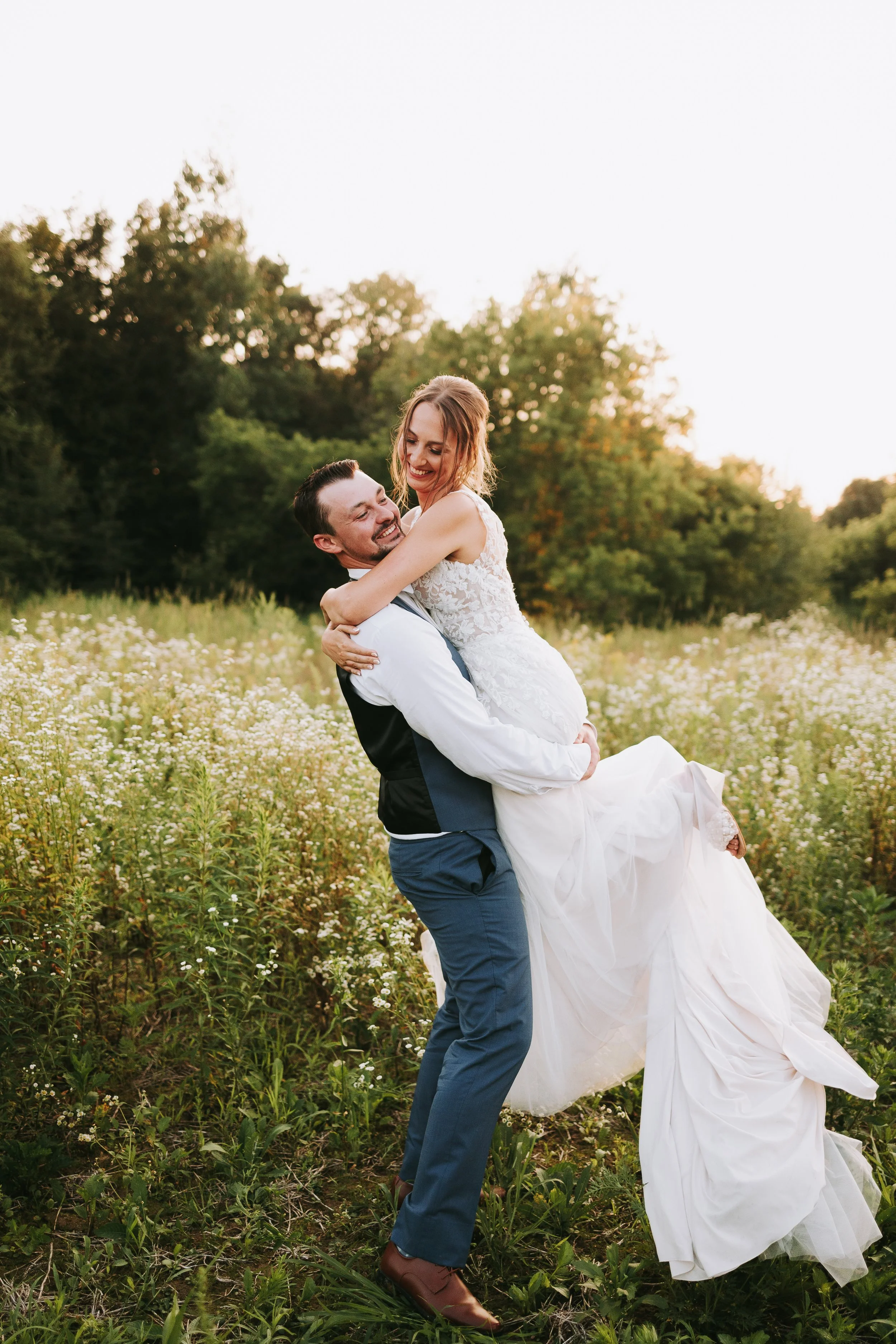 A happy couple in wedding attire, with the groom lifting the bride in a field of wildflowers during sunset.