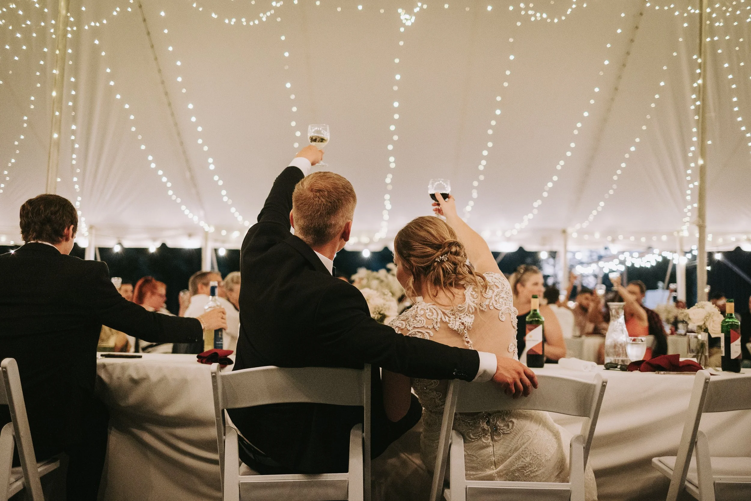 A wedding reception with guests raising glasses in toast under a decorated tent with string lights.