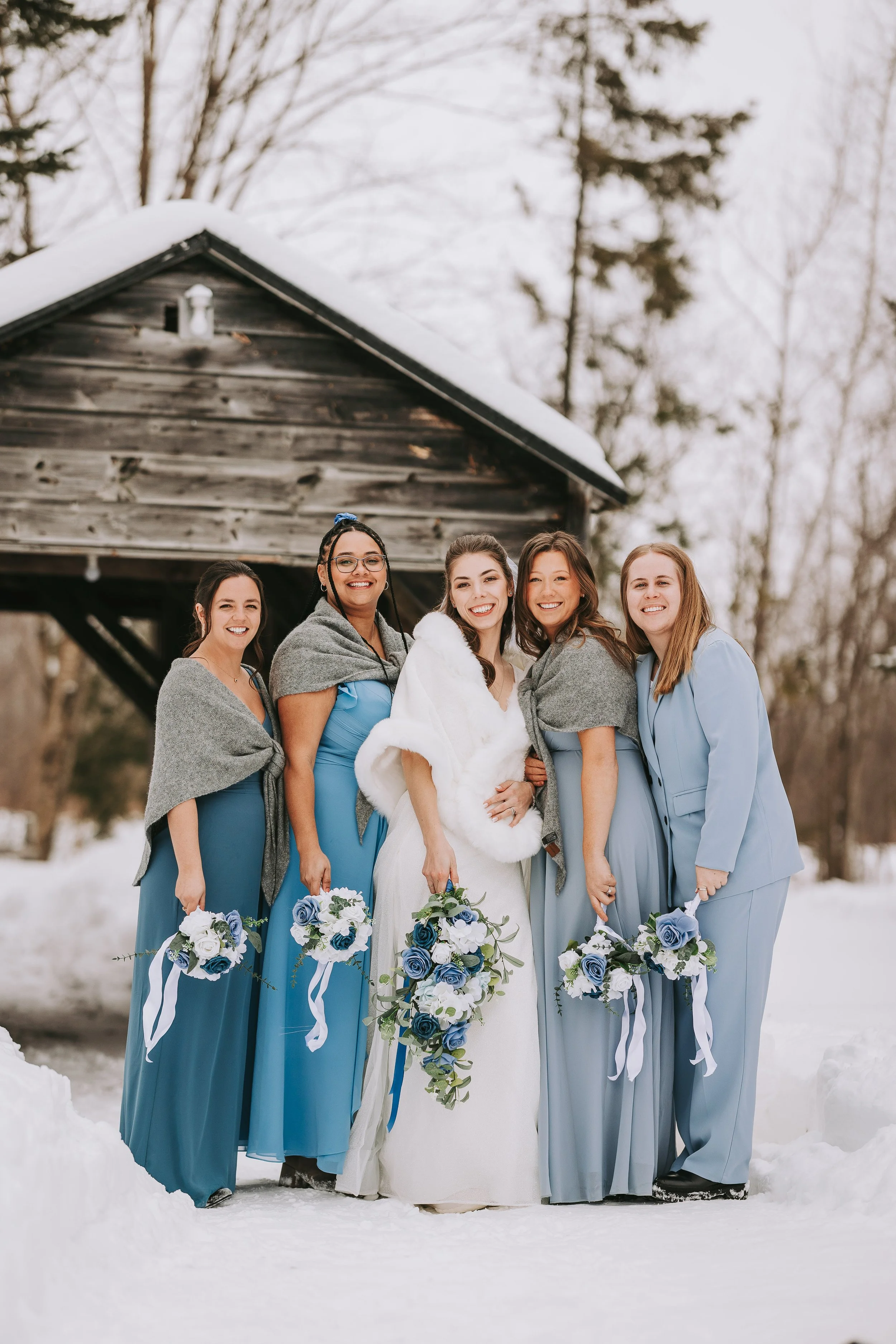 Group of women in blue dresses and gray shawls, with one woman in a white wedding dress, standing in snow outdoors, holding bouquets, smiling.