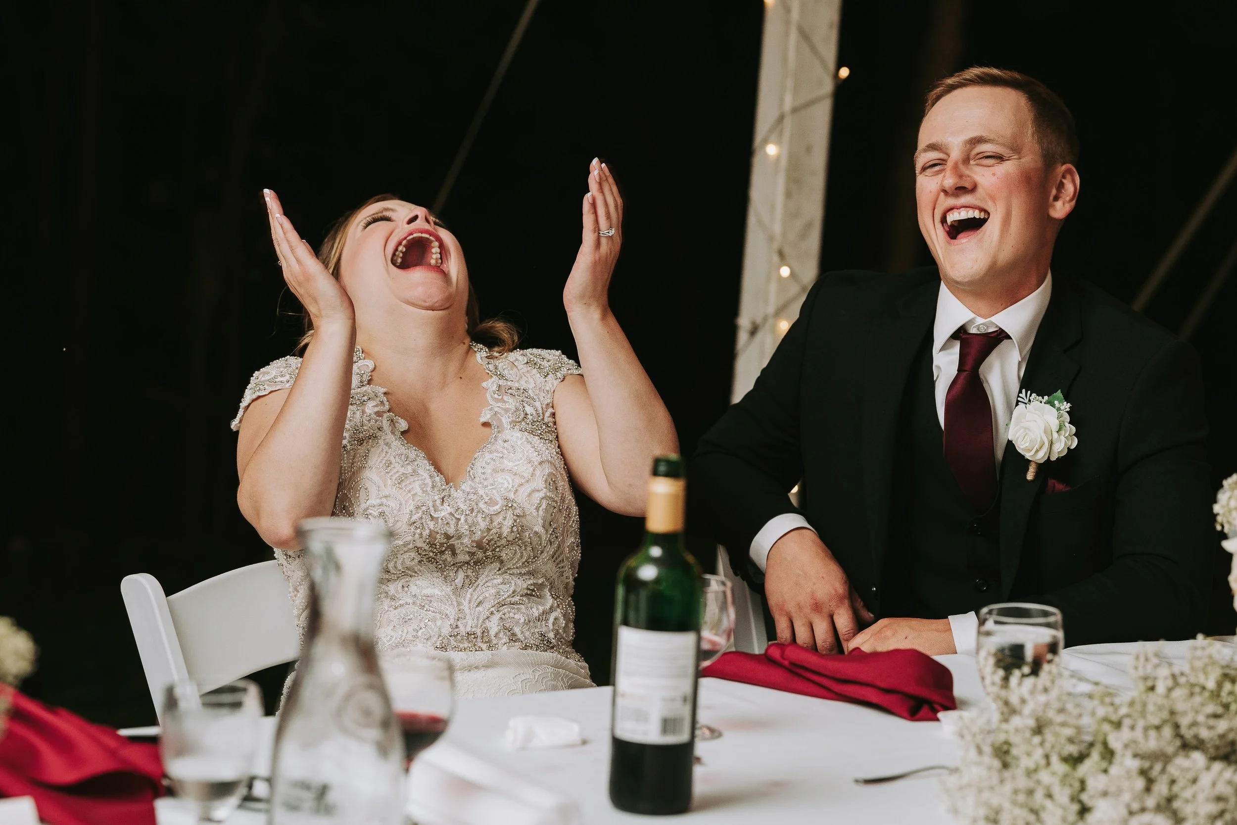A bride and groom at a wedding reception are laughing joyfully, with the bride tilting her head back and the groom smiling widely.