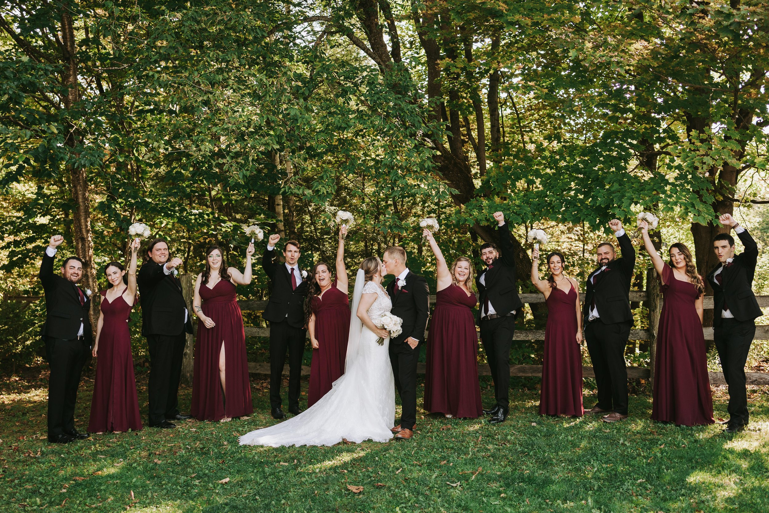 Wedding party standing outdoors with trees in the background, the bride and groom in the center sharing a kiss, surrounded by bridesmaids and groomsmen celebrating with raised fists, some holding bouquets.