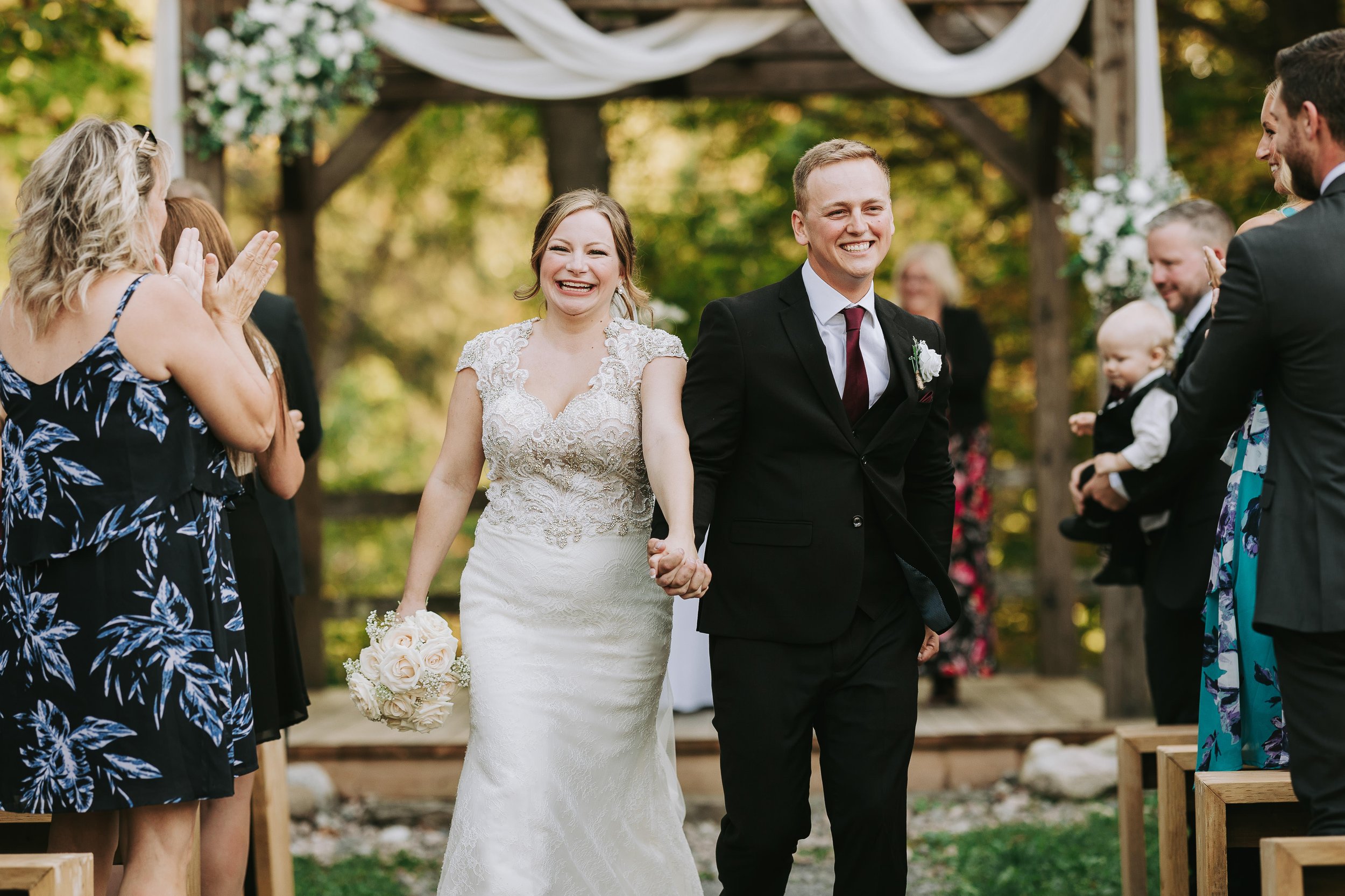 A newlywed couple walking down the aisle at their outdoor wedding ceremony, holding hands and smiling. The bride is wearing a lace wedding dress and holding a bouquet of roses, while the groom is dressed in a black suit with a white shirt and maroon 