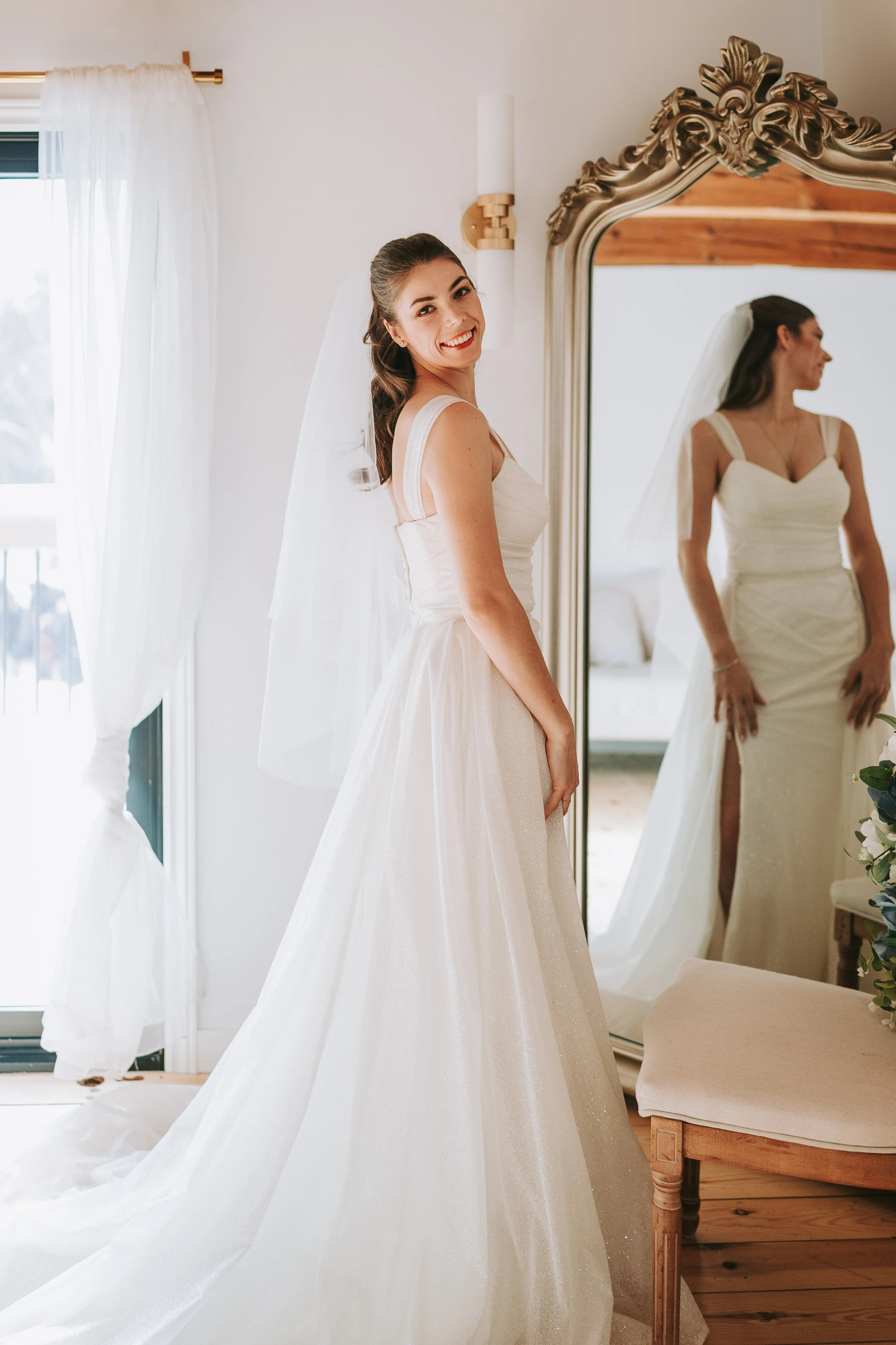 A smiling bride in a white wedding dress standing in front of a large mirror, adjusting her dress, with a window and sheer curtains in the background.