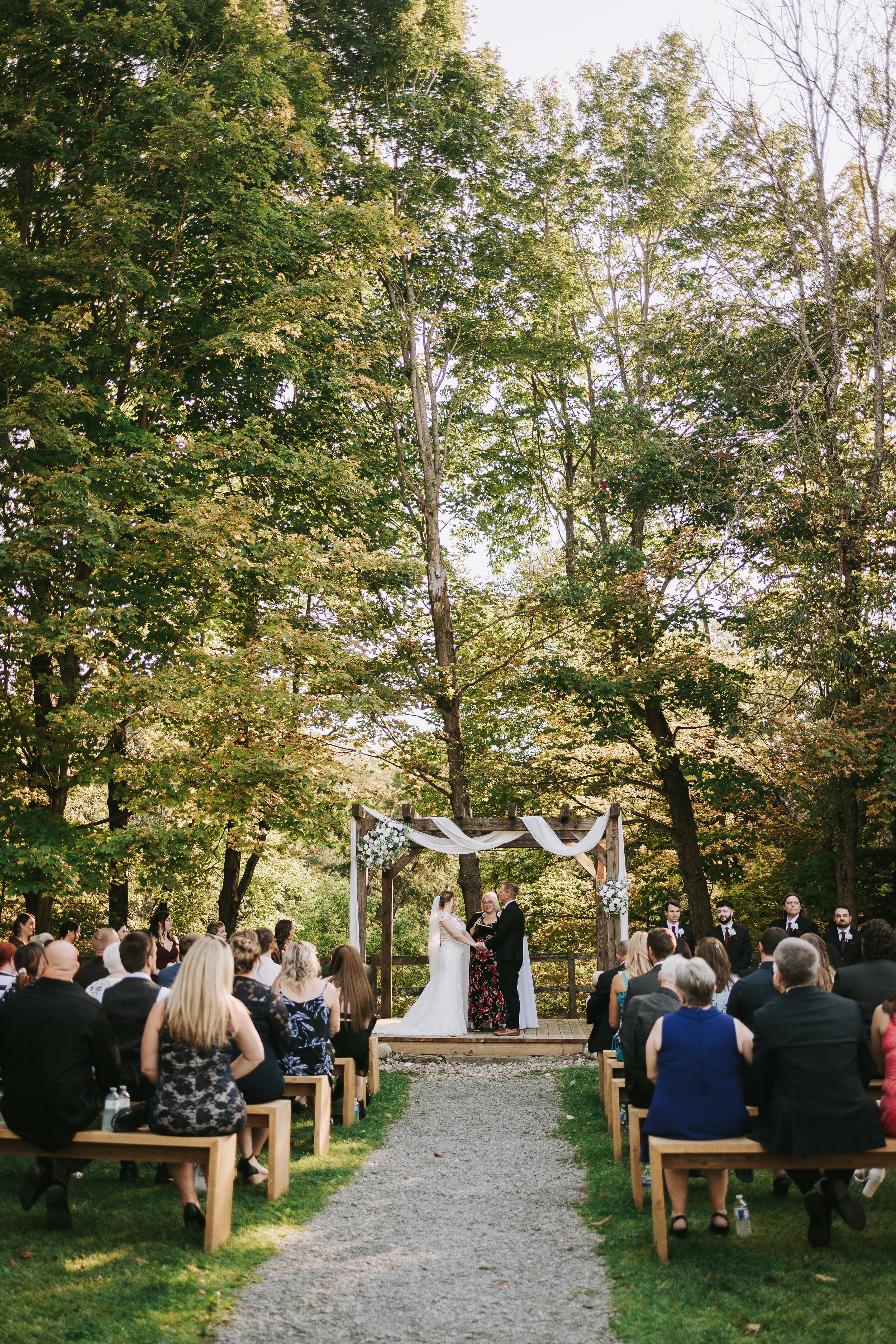 A wedding ceremony outdoors on a wooden platform under a decorated arch. The bride and groom are exchanging vows, surrounded by seated guests in a lush, green, wooded setting.