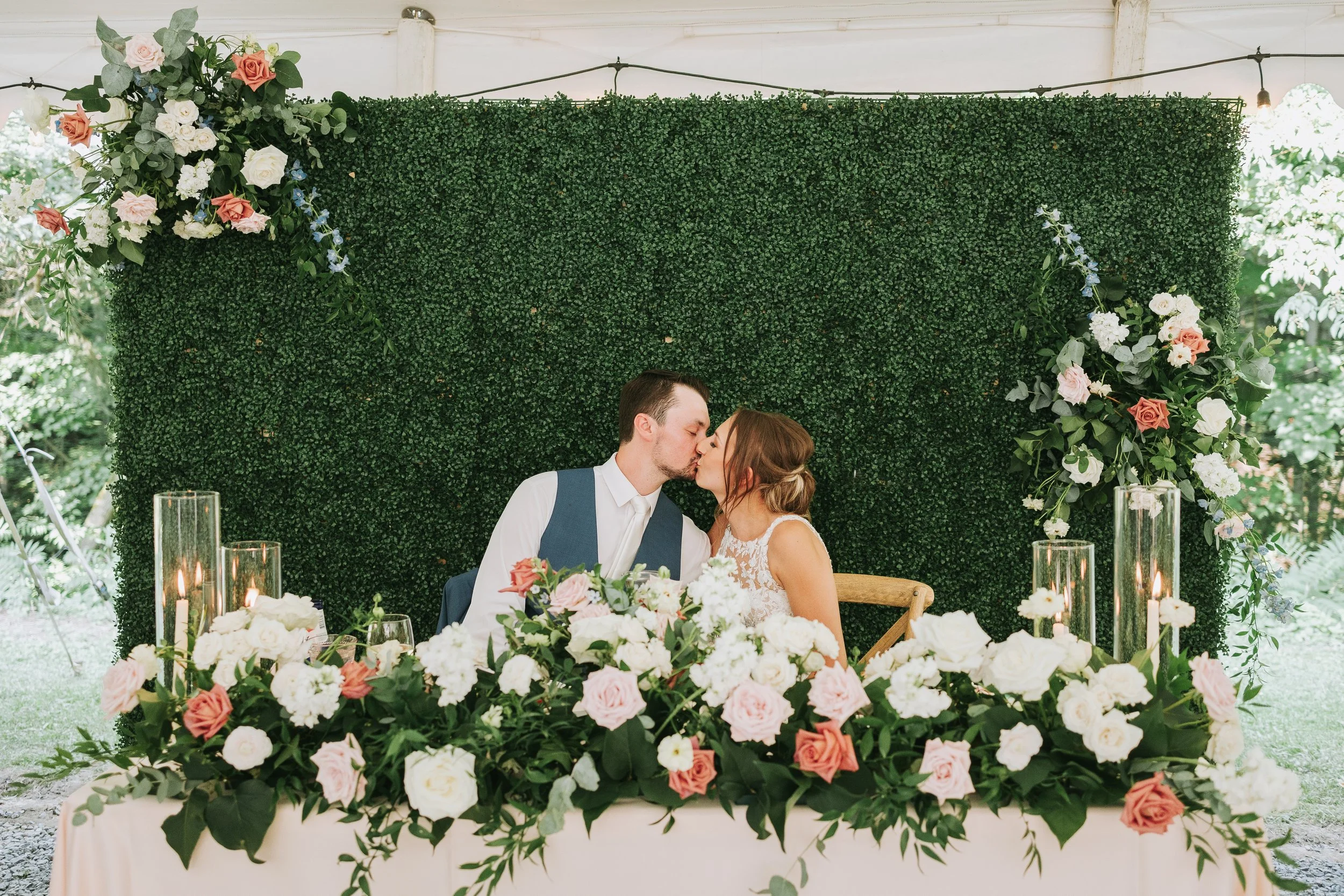 A newlywed couple sharing a kiss at their wedding reception table decorated with a lush floral arrangement, with a green hedge backdrop and floral accents on either side.