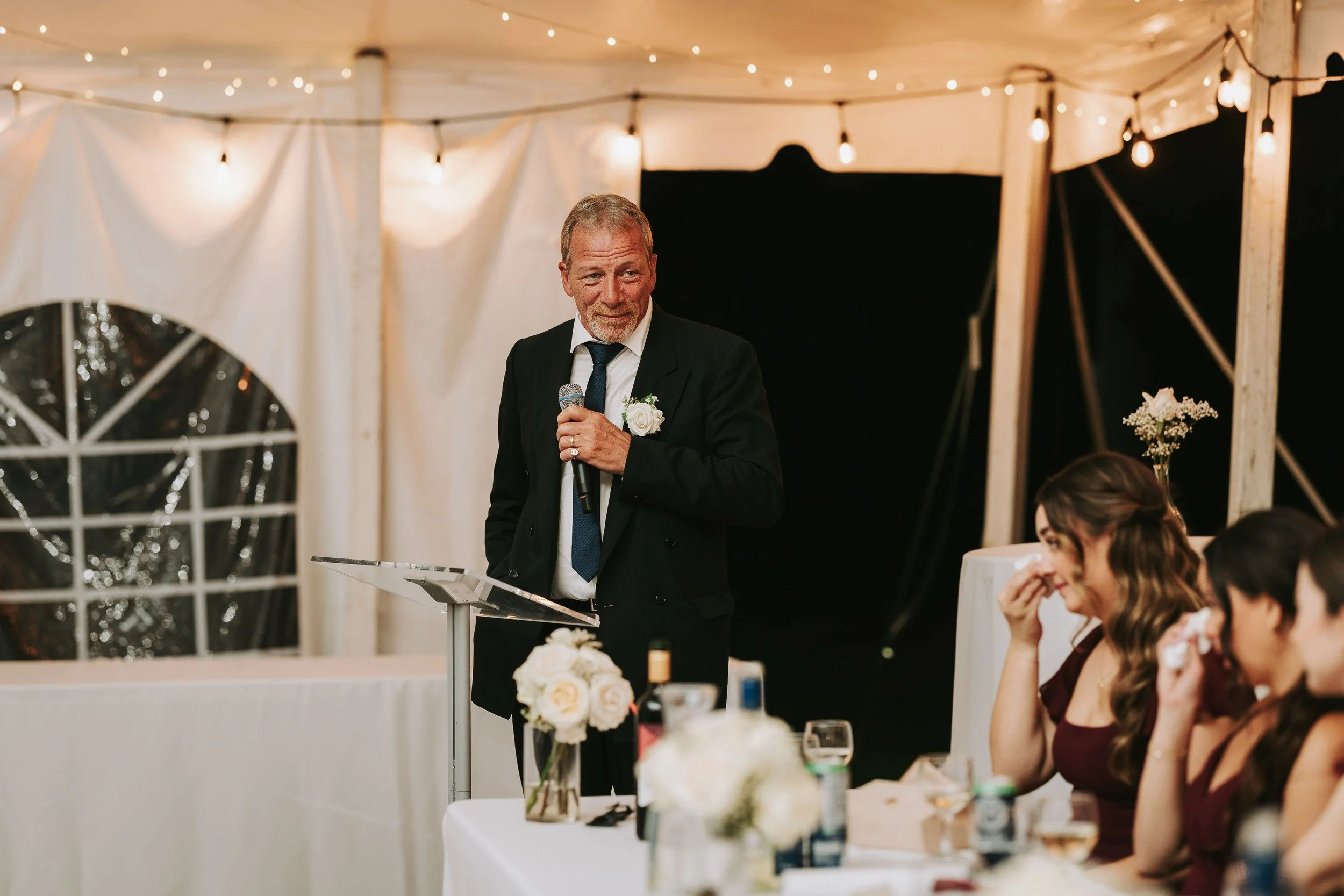 Older man in a suit giving a speech at a wedding reception, holding a microphone, with women seated at a table laughing and smiling.