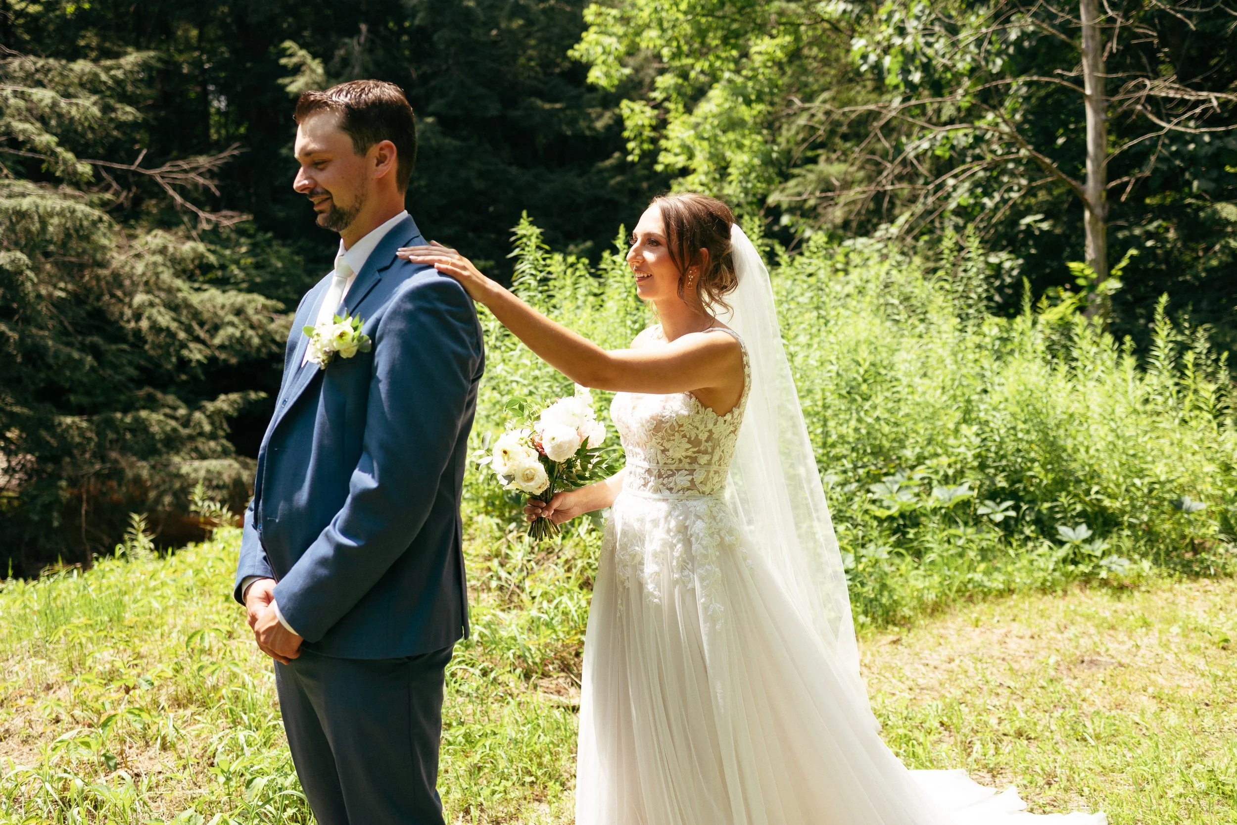 Bride placing hand on groom's shoulder during outdoor wedding in a wooded area.