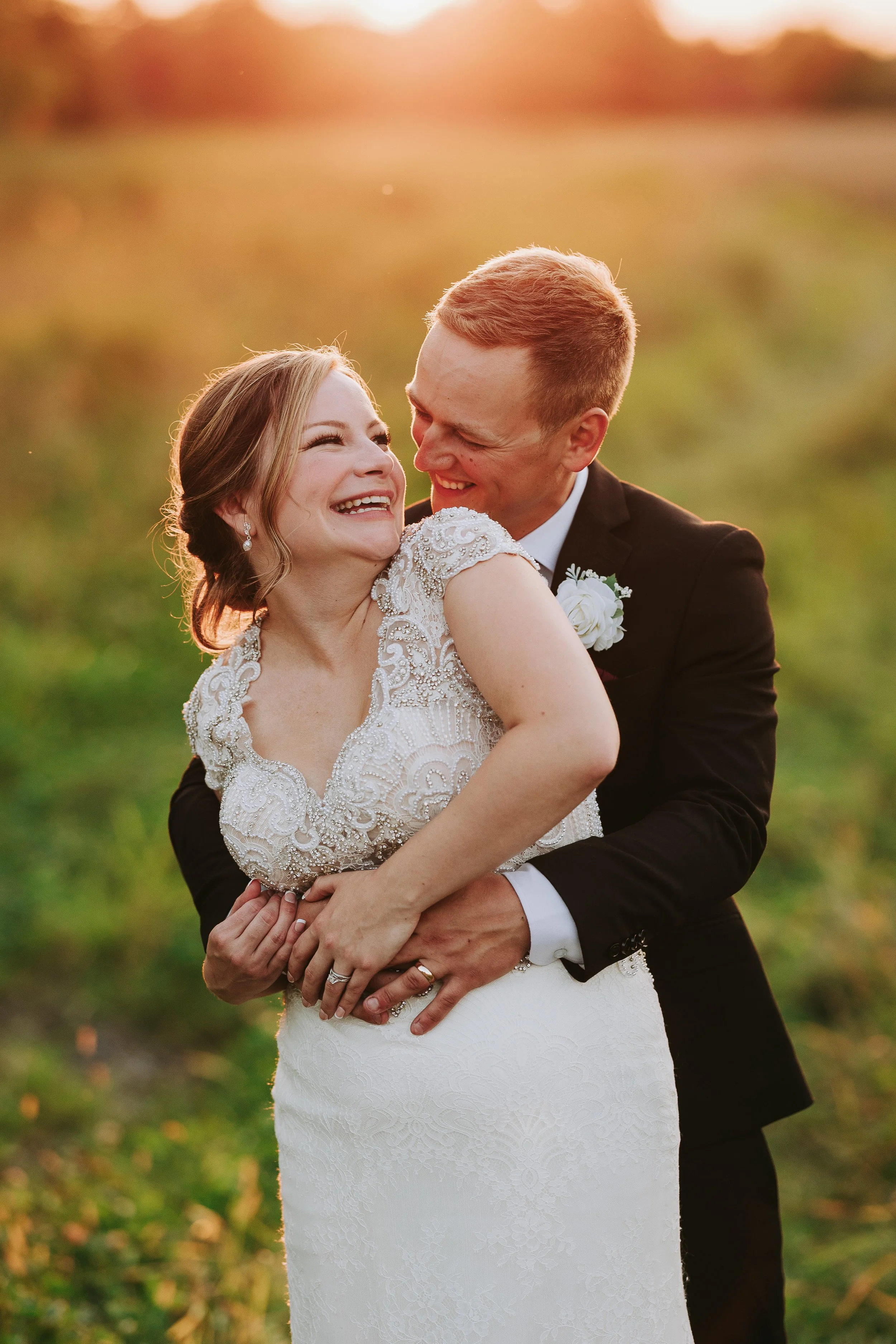 A newlywed couple embracing happily outdoors during sunset, with the groom in a black tuxedo and the bride in a white lace wedding dress, smiling at each other.