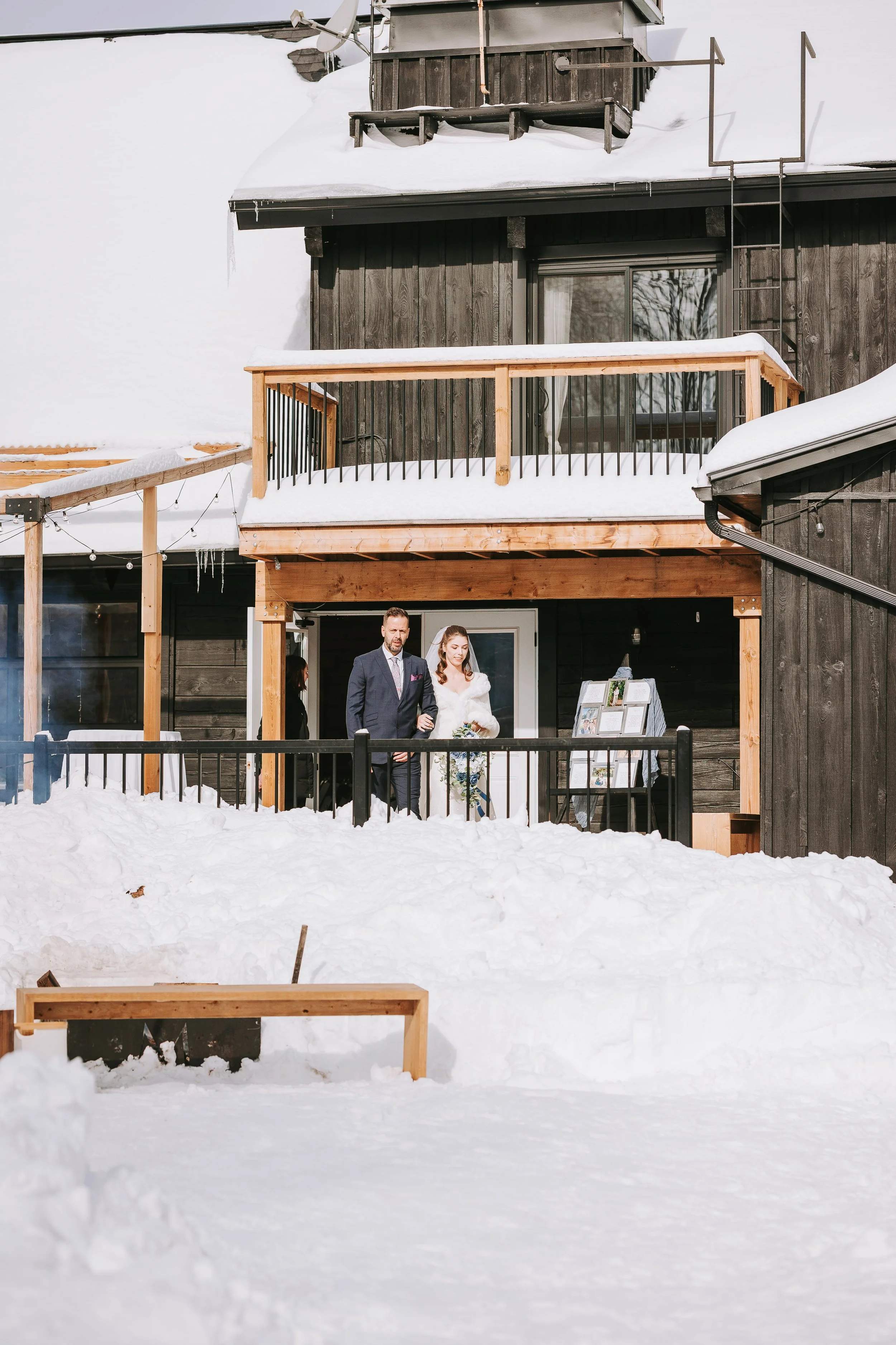 A couple dressed in wedding attire walking out onto a snowy patio of a dark wood house with a new balcony, while snow covers the ground in front of the house.