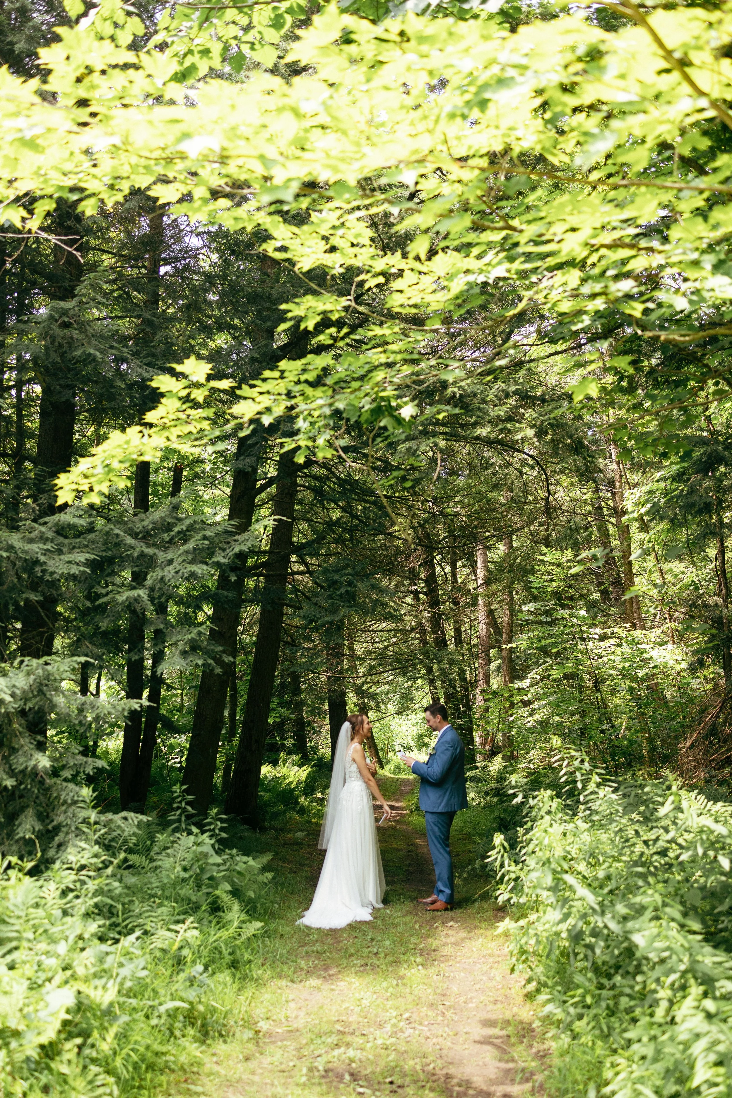 A bride and groom standing on a forest path during their wedding, surrounded by lush green trees and foliage.