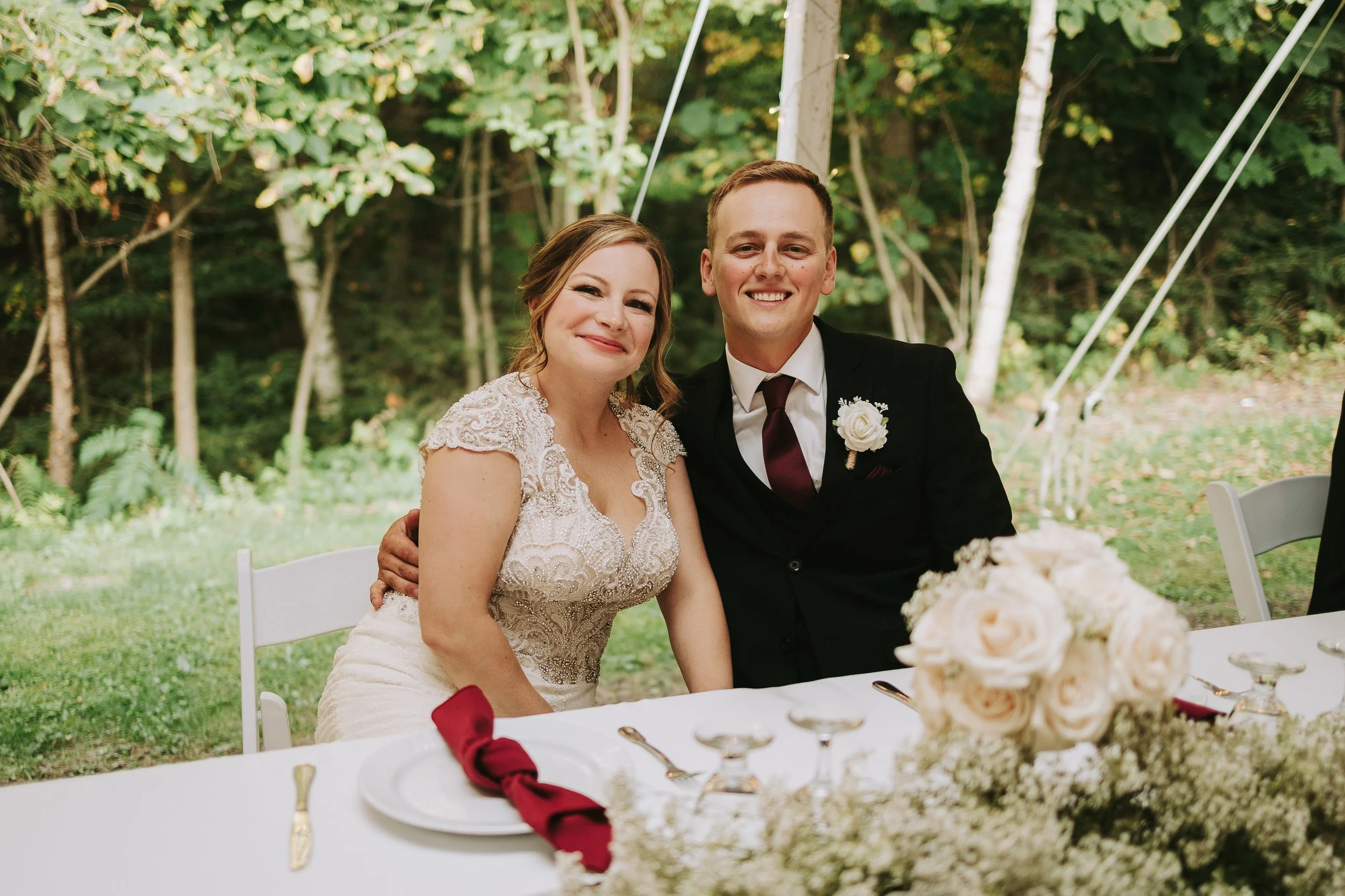 A bride and groom sitting together outdoors at a wedding reception, smiling at the camera, with a table set with flowers, plates, and cutlery in front of them.