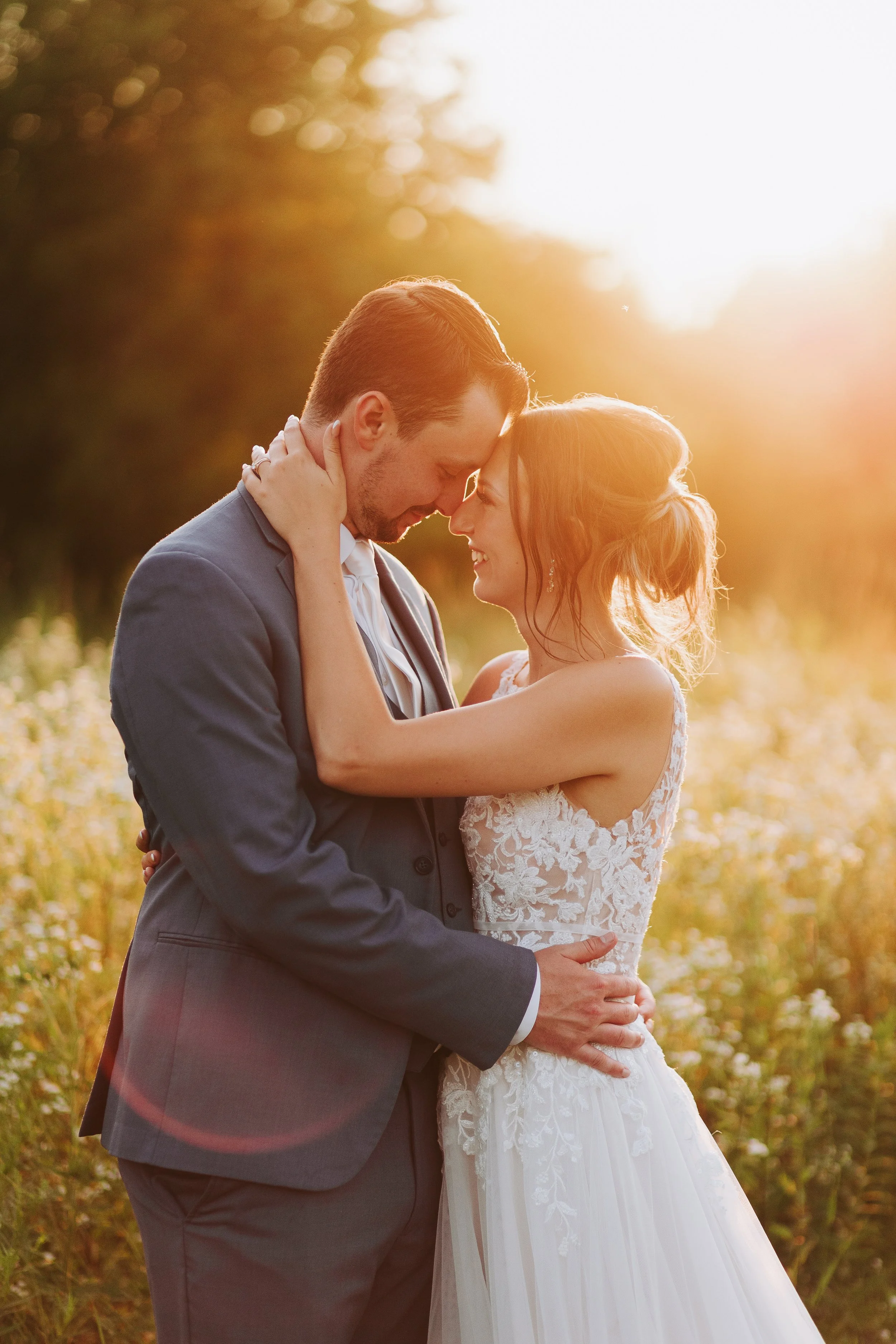 A bride and groom embrace each other outdoors during sunset, touching foreheads and smiling.