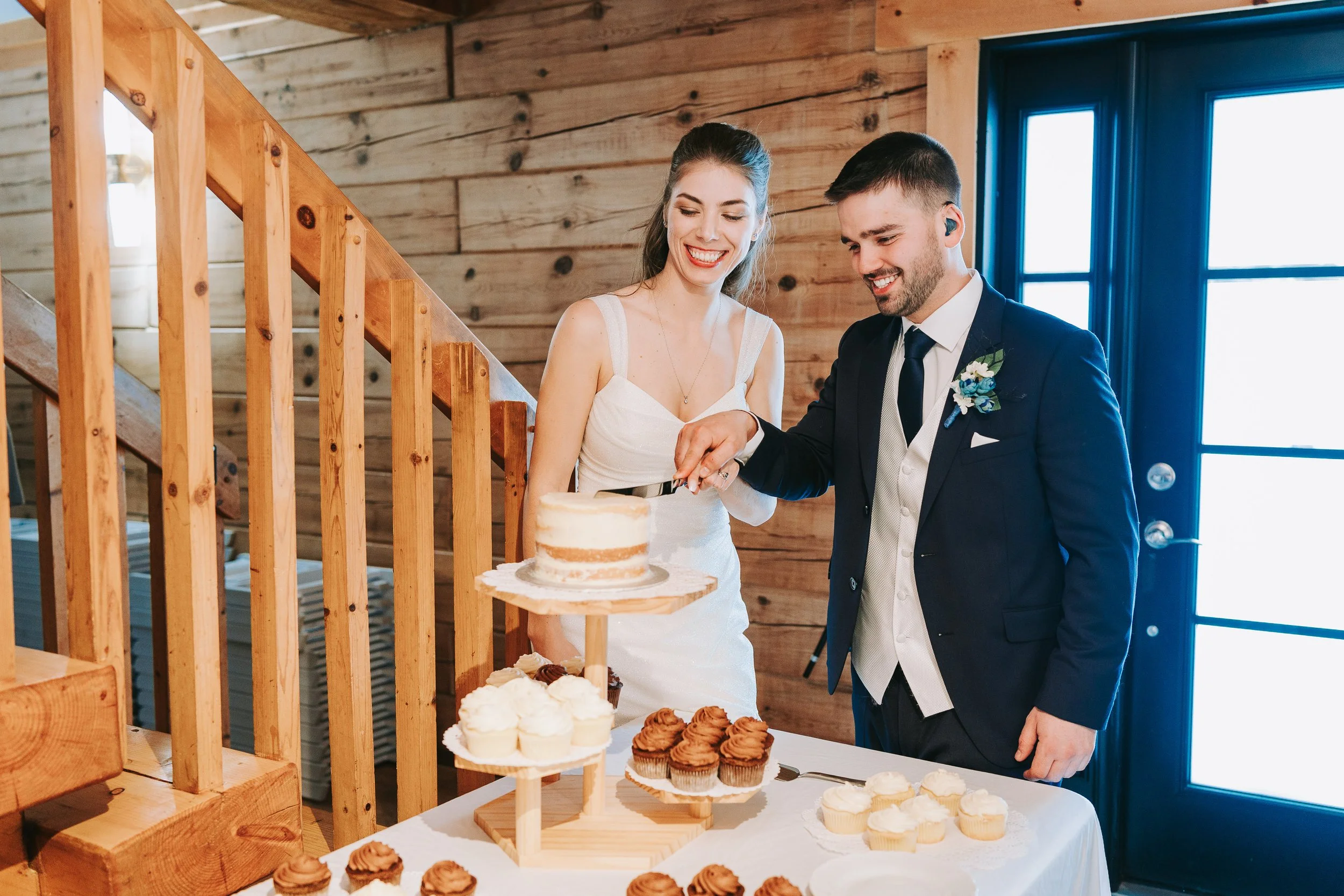 A bride and groom in formal wedding attire cutting a cake at their wedding reception, with desserts on a table in a beautiful Ottawa venue.