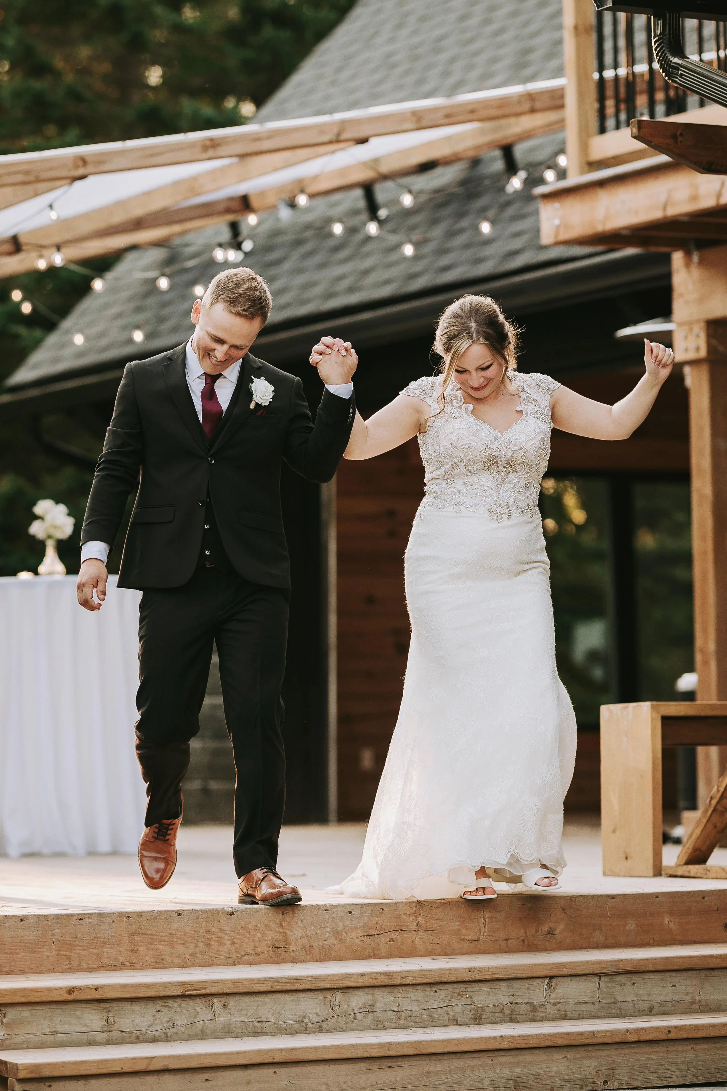 A newlywed couple dancing outdoors on a wooden platform, holding hands with arms raised, surrounded by string lights and a rustic wooden structure, during sunset.