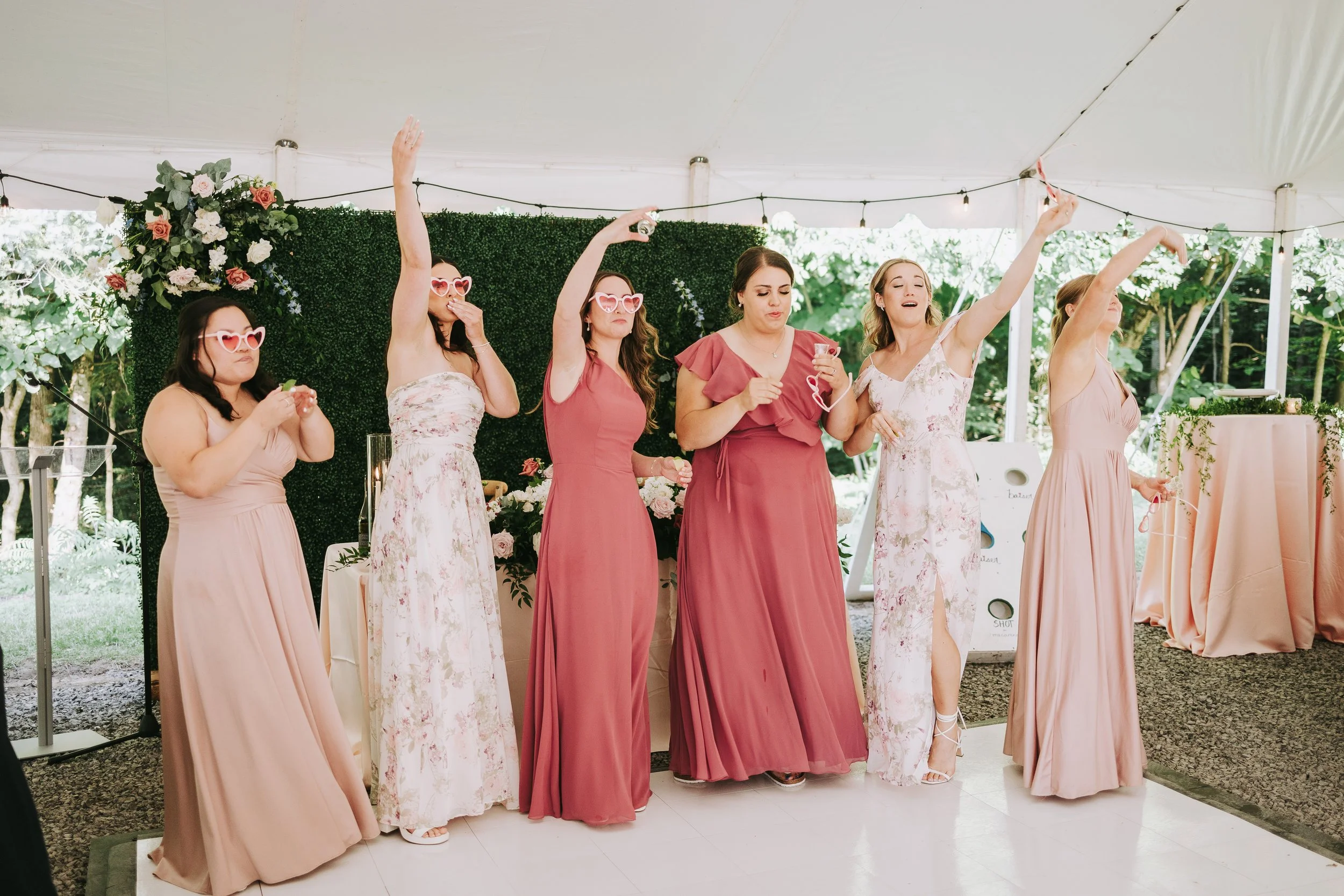 Group of six women in pink and floral dresses dancing and celebrating at a wedding reception under a tent with greenery background.