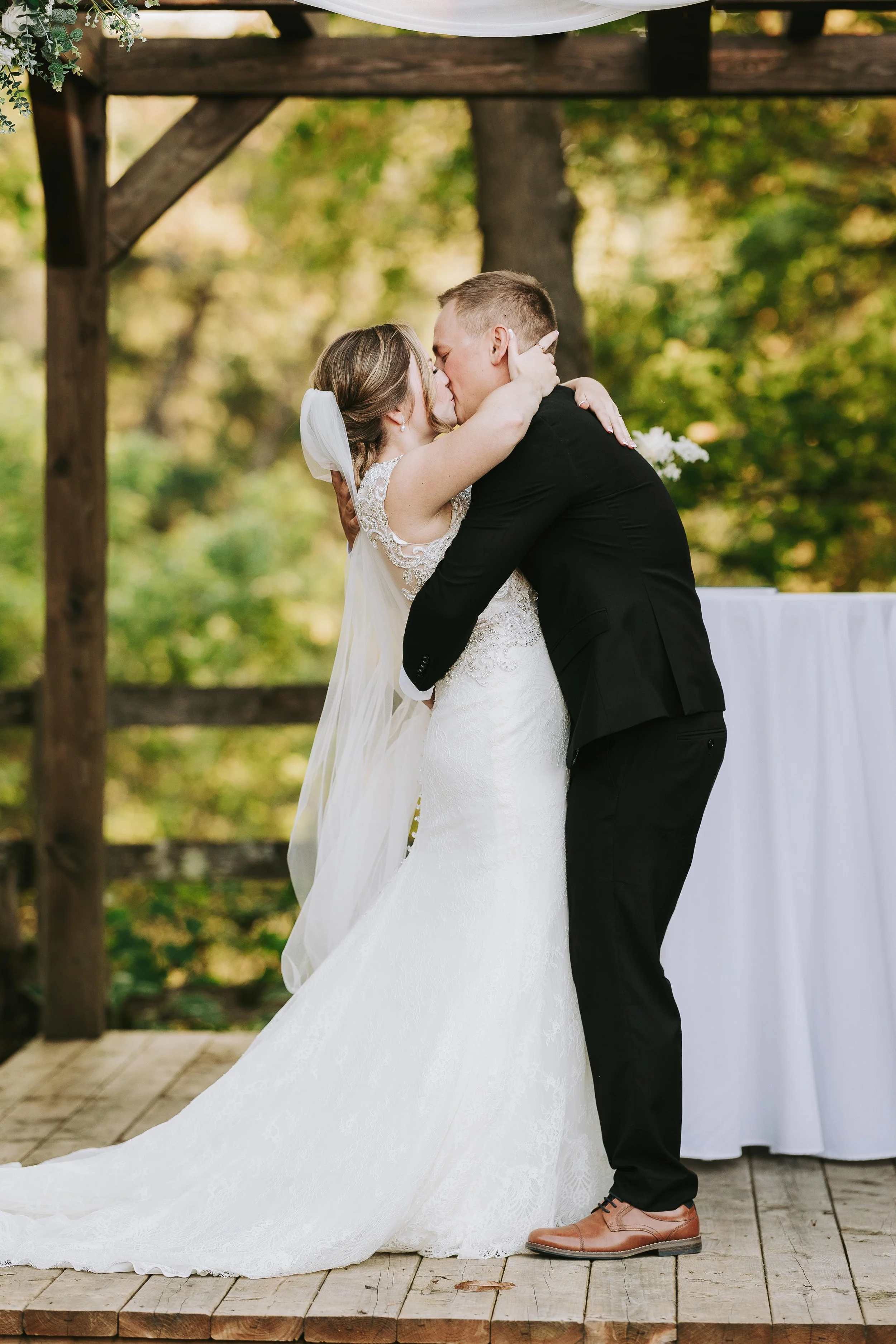 A bride and groom kiss during their wedding ceremony outdoors on a wooden platform, surrounded by greenery.