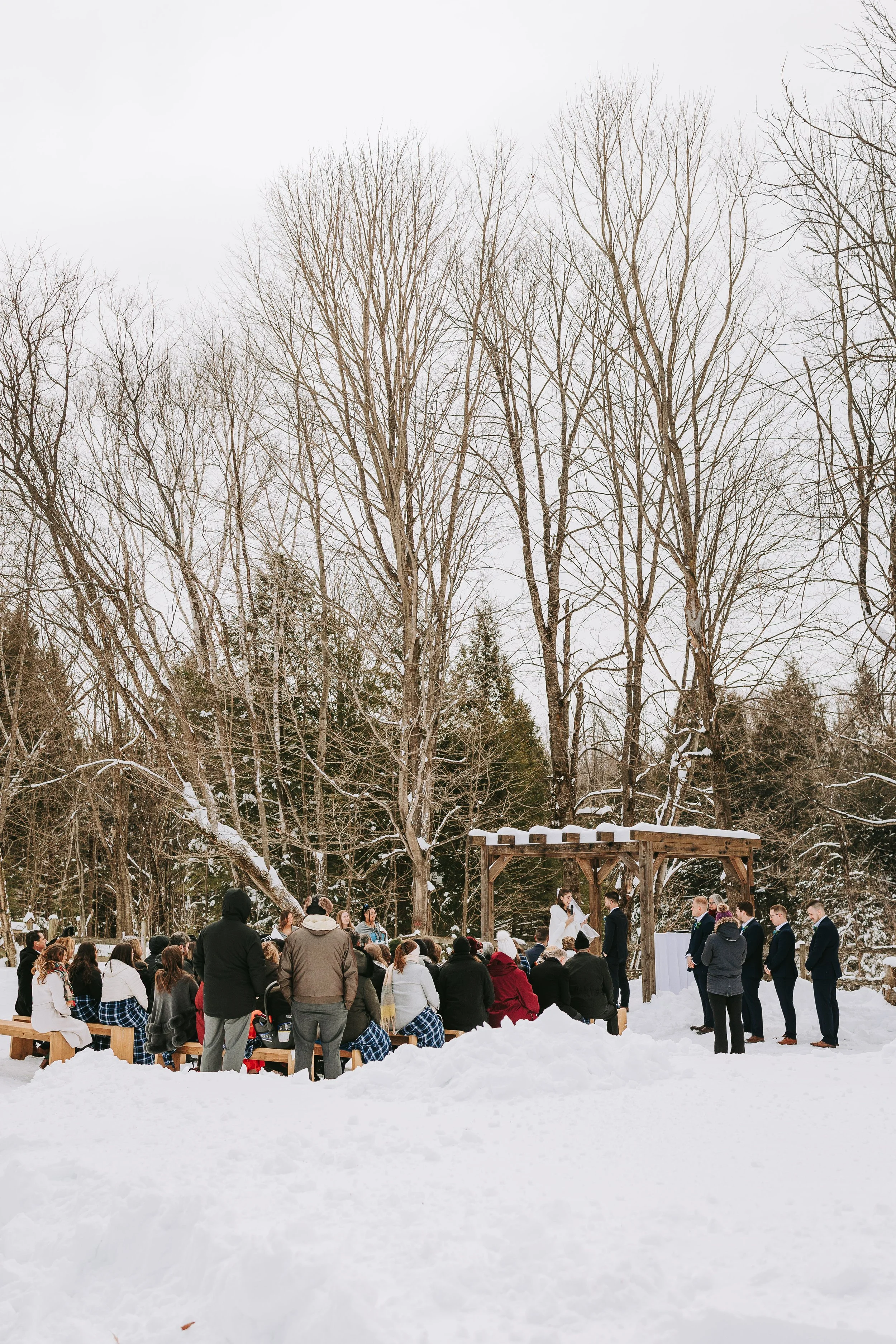 Wedding ceremony happening outdoors in snowy winter, with guests seated and standing under a wooden pergola, trees in the background.