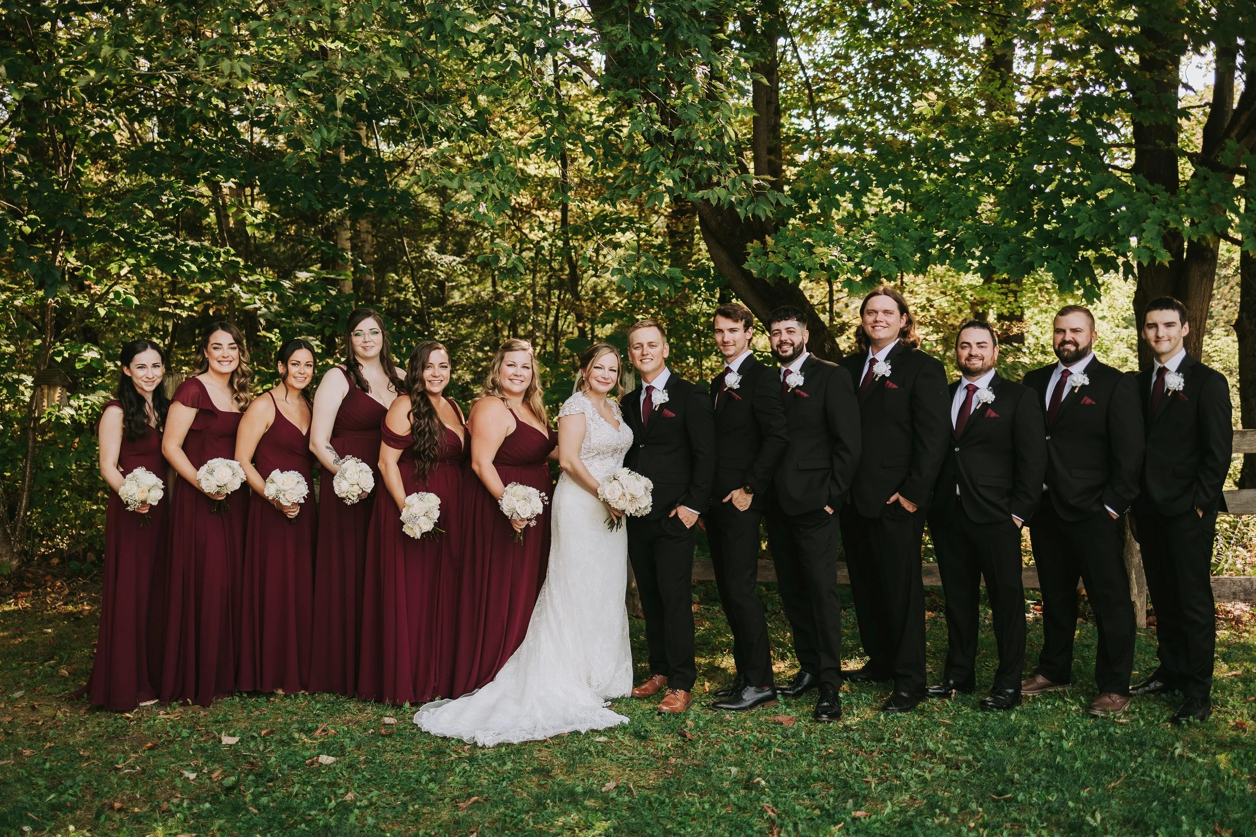 A wedding party outdoors, with bridesmaids in burgundy dresses holding bouquets and groomsmen in black suits with burgundy ties, standing in front of trees and greenery.