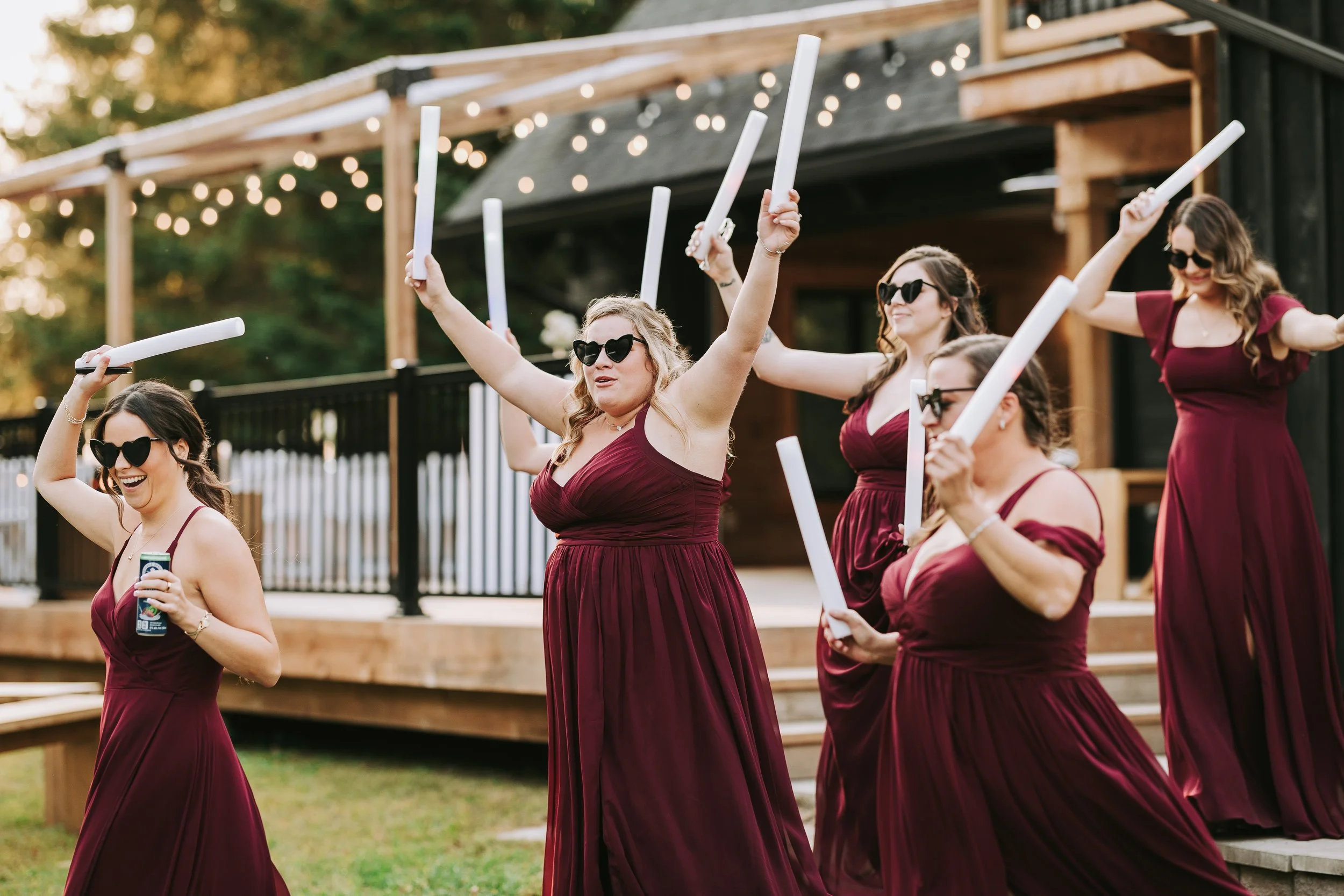 Group of women in matching burgundy dresses celebrating outdoors at sunset, holding glowing sticks, and wearing sunglasses.