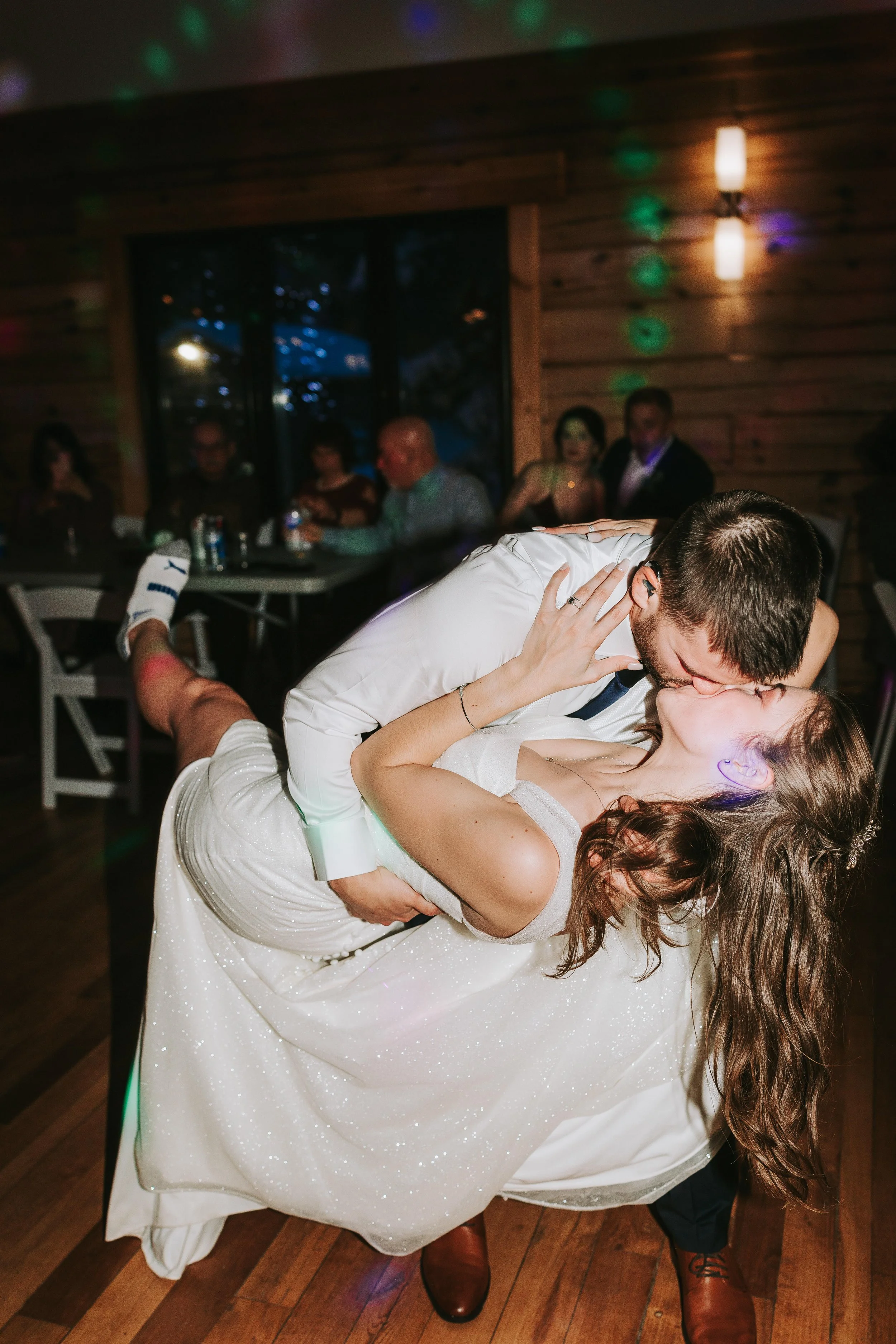 A couple kissing and dancing at a wedding reception, with guests seated at tables in a rustic wooden room with dim lighting.