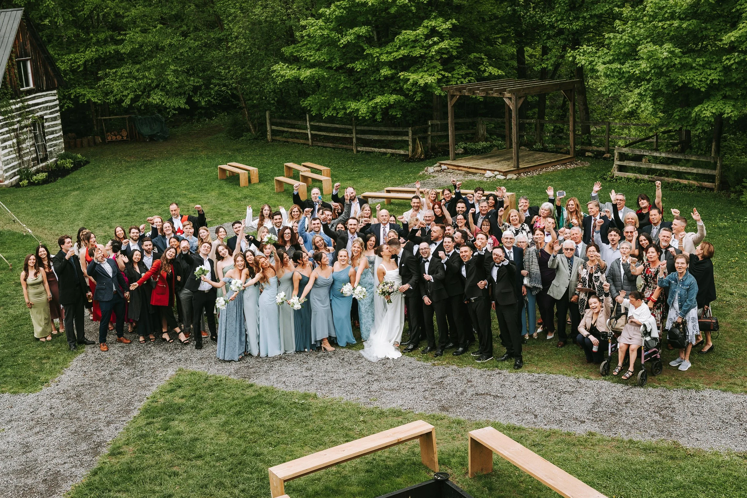 A large group of people gathered outdoors on a grassy area for a wedding celebration, with trees and wooden structures in the background.