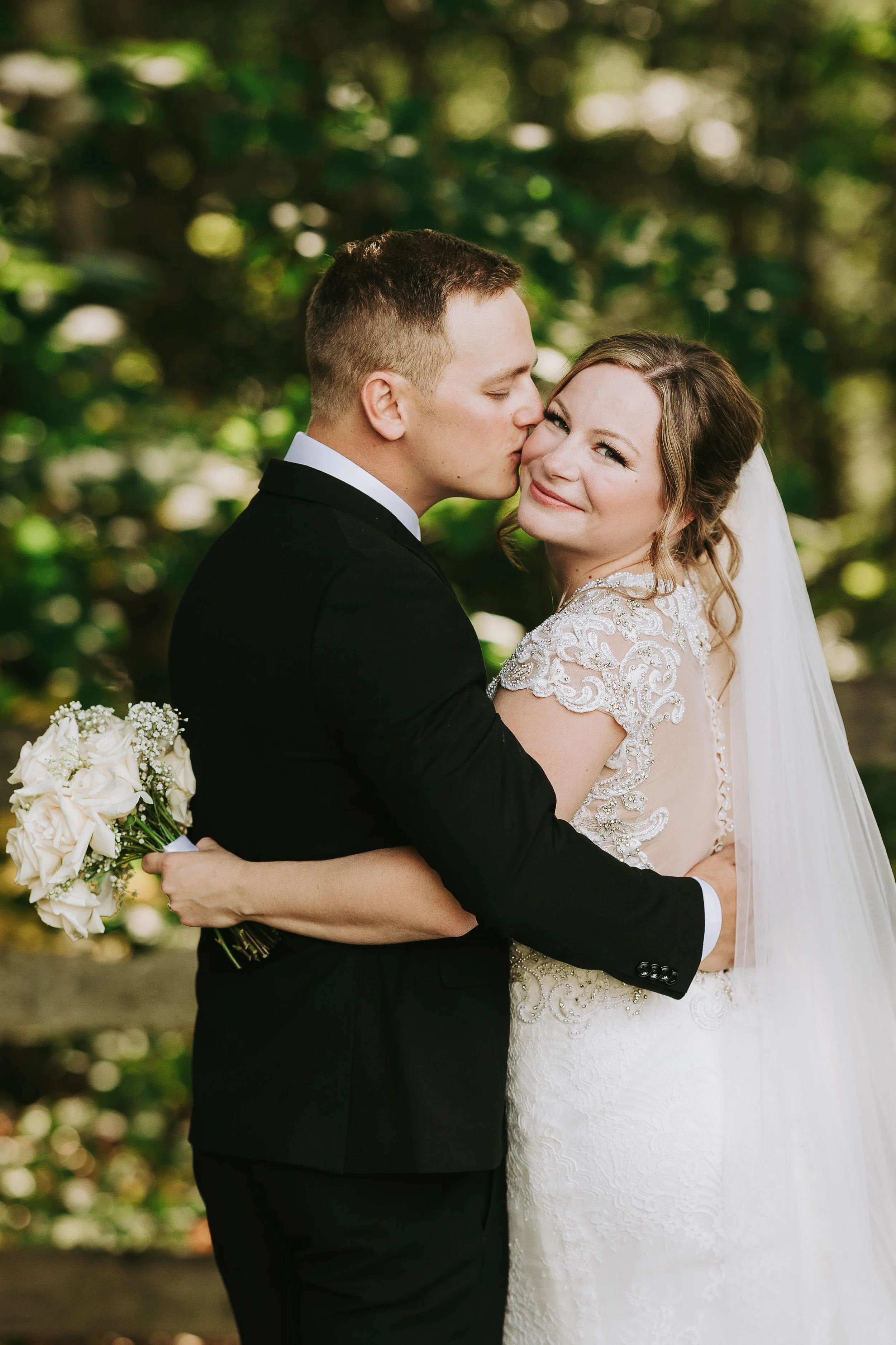 A bride and groom embrace outdoors, the groom kisses the bride on the cheek as she smiles with her eyes closed, holding a bouquet of white roses.