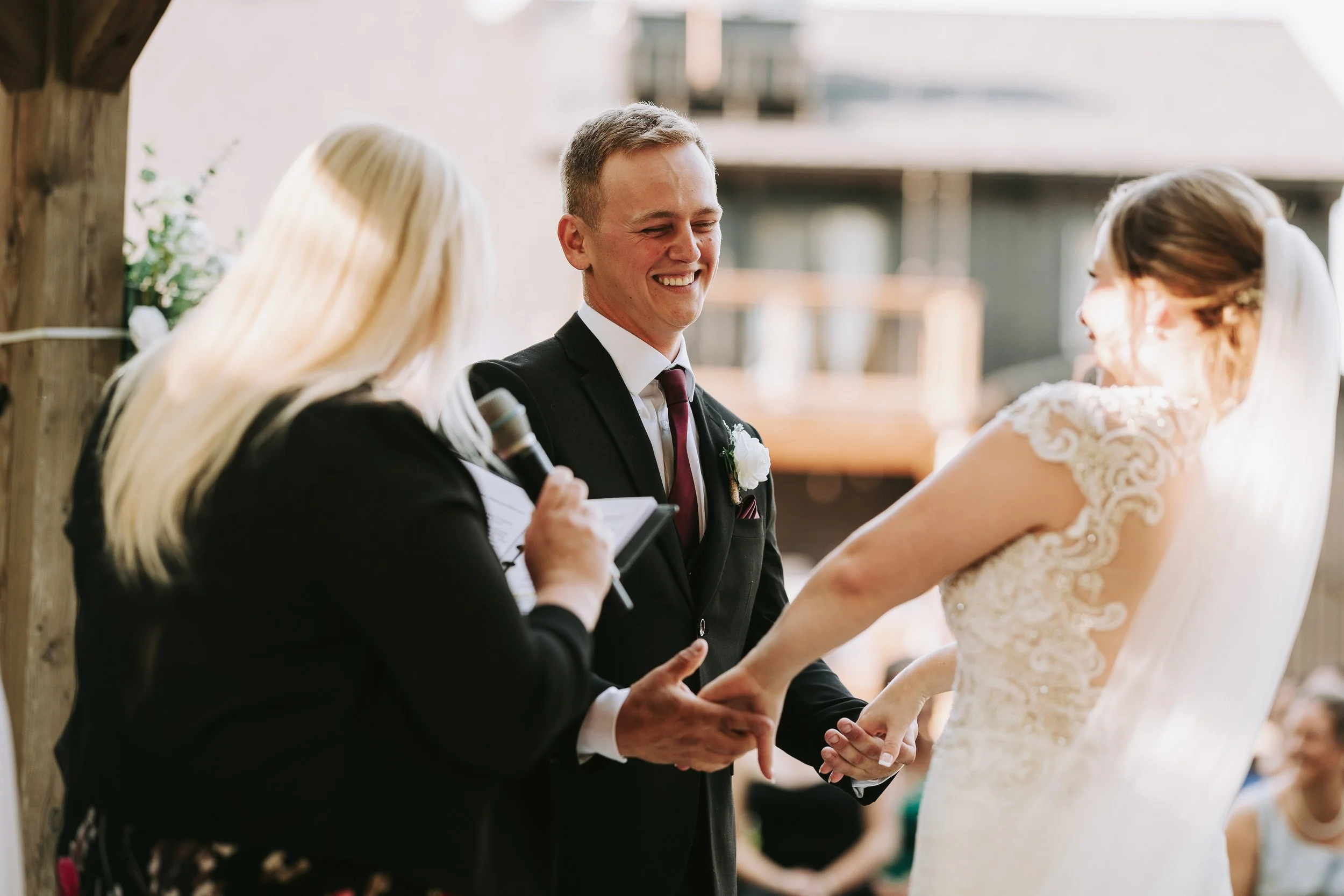 A wedding ceremony with a smiling groom holding hands with the bride as she laughs during their vows, an officiant reading from a paper, in an outdoor setting with friends in the background.