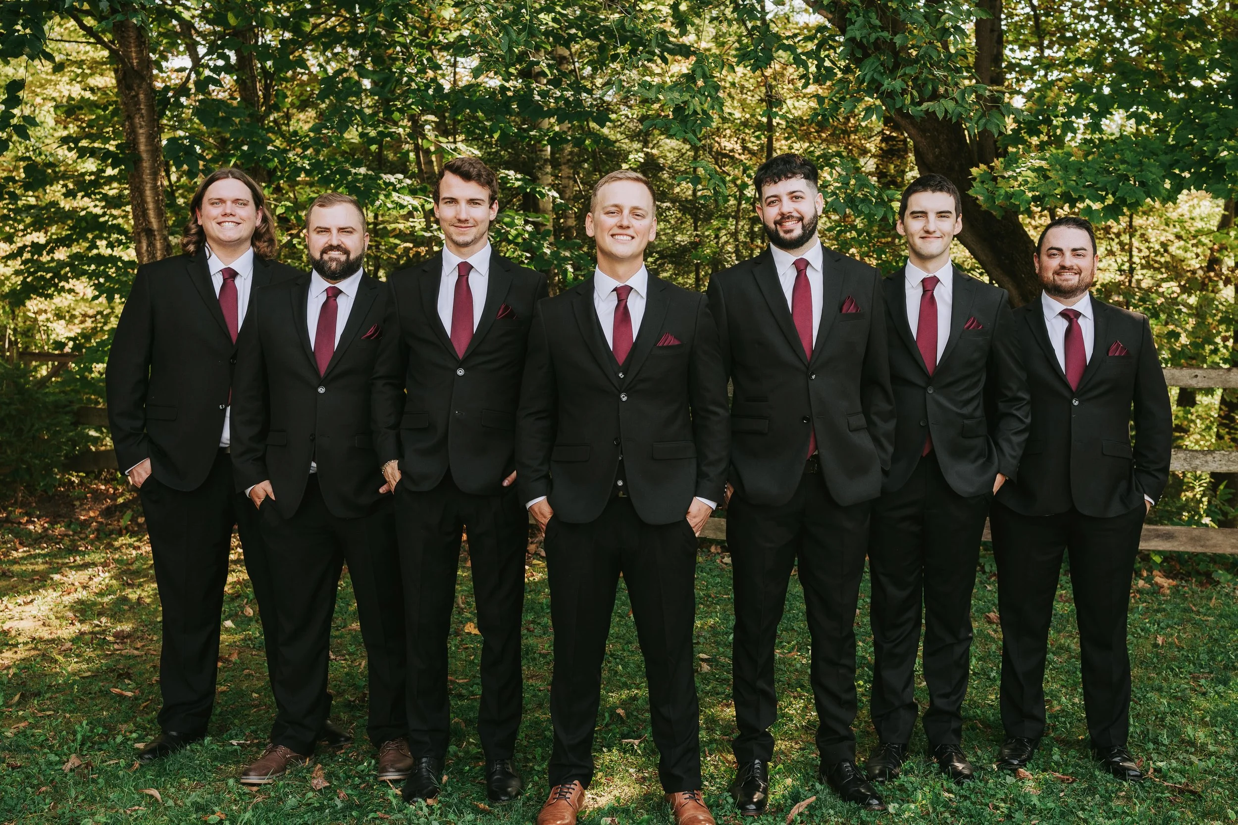 Group of seven men in black suits, white shirts, and maroon ties standing outdoors on grass with green trees in background.