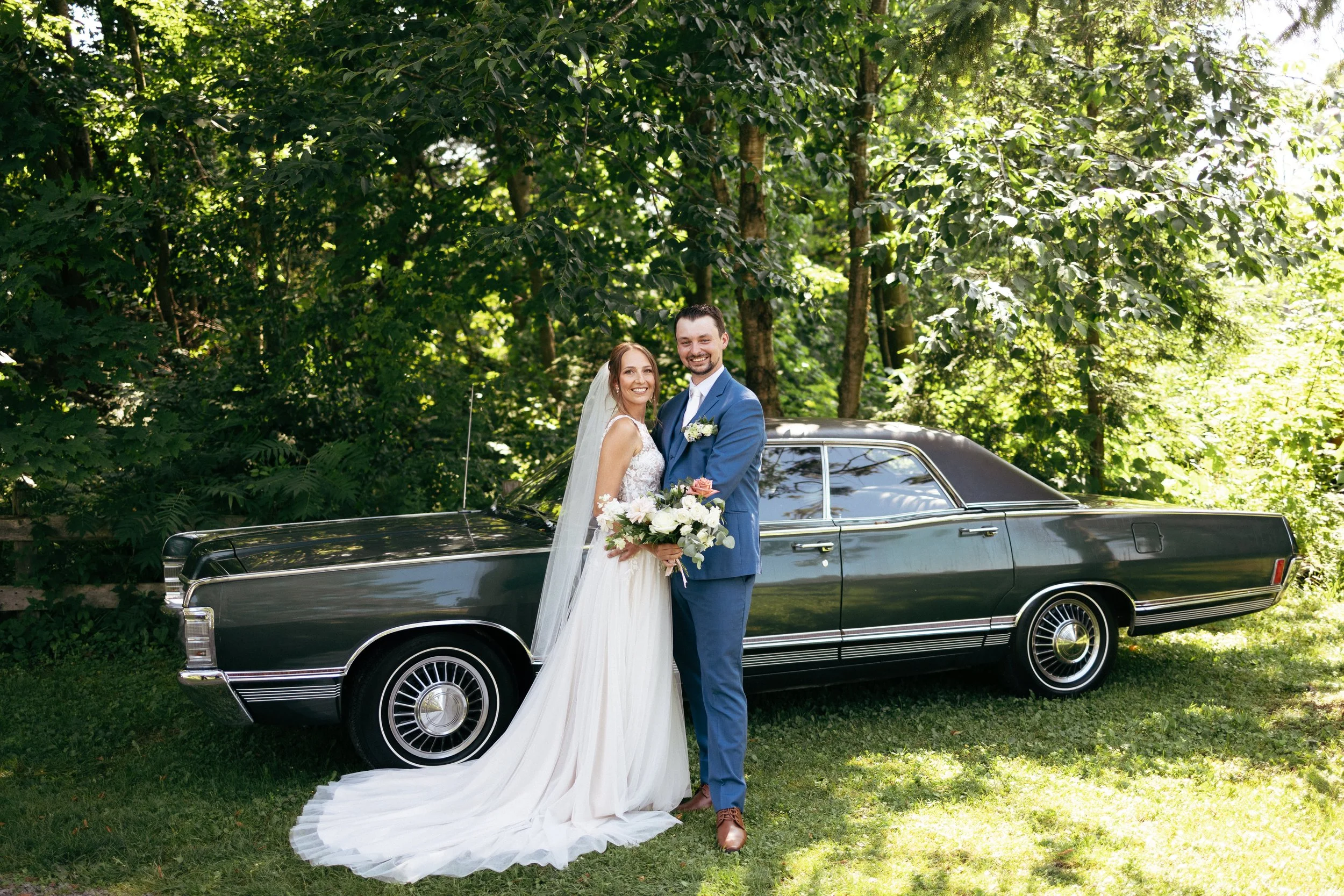 A newlywed couple standing outdoors in front of a black vintage car and surrounded by lush green trees, smiling at the camera. The bride wears a white wedding dress and veil, holding a bouquet of flowers. The groom wears a blue suit with a white shir