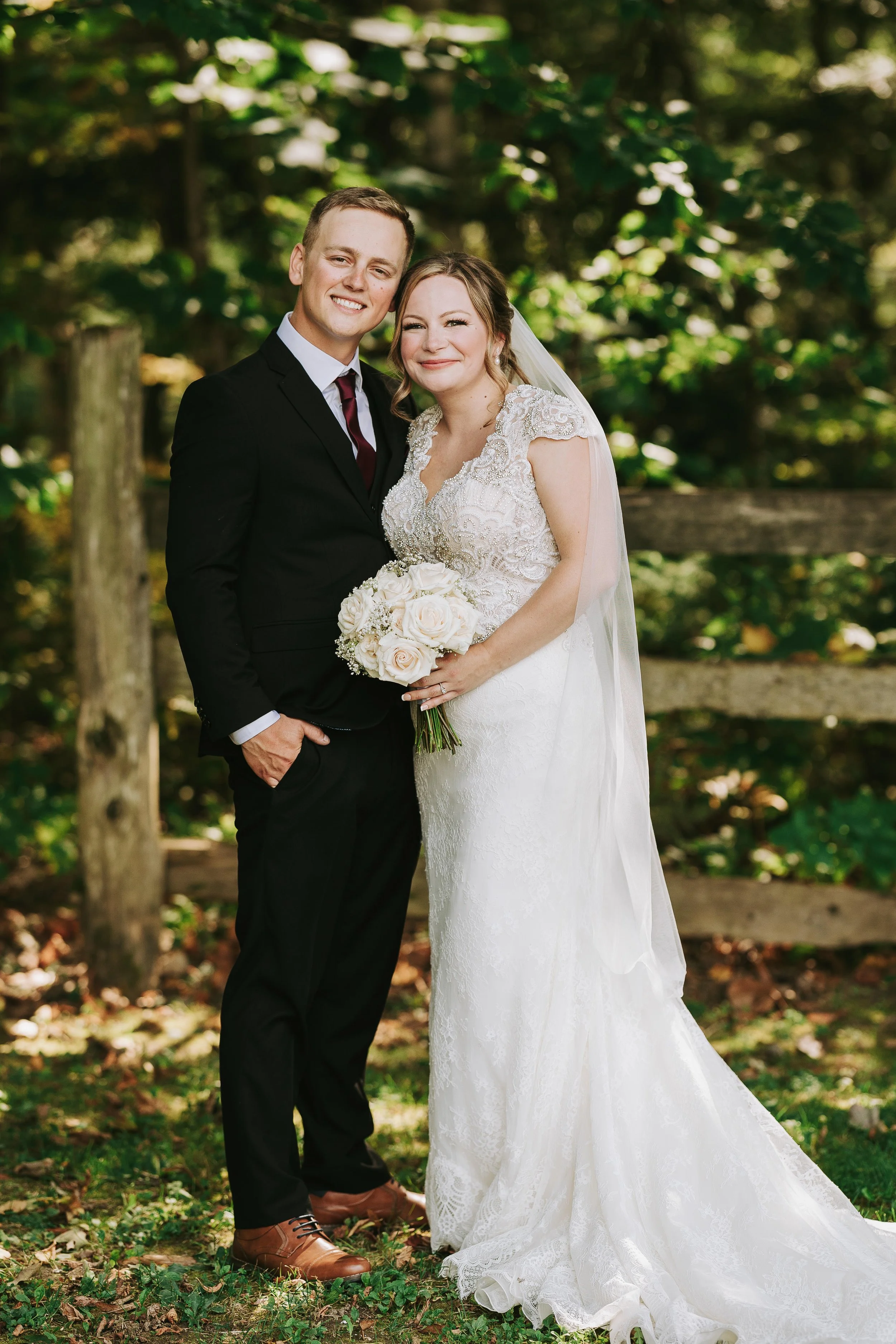 A newlywed couple standing outdoors in a wooded area, smiling at the camera. The bride is wearing a white lace wedding gown and holding a bouquet of white roses, while the groom is in a black suit with a white shirt and dark tie.