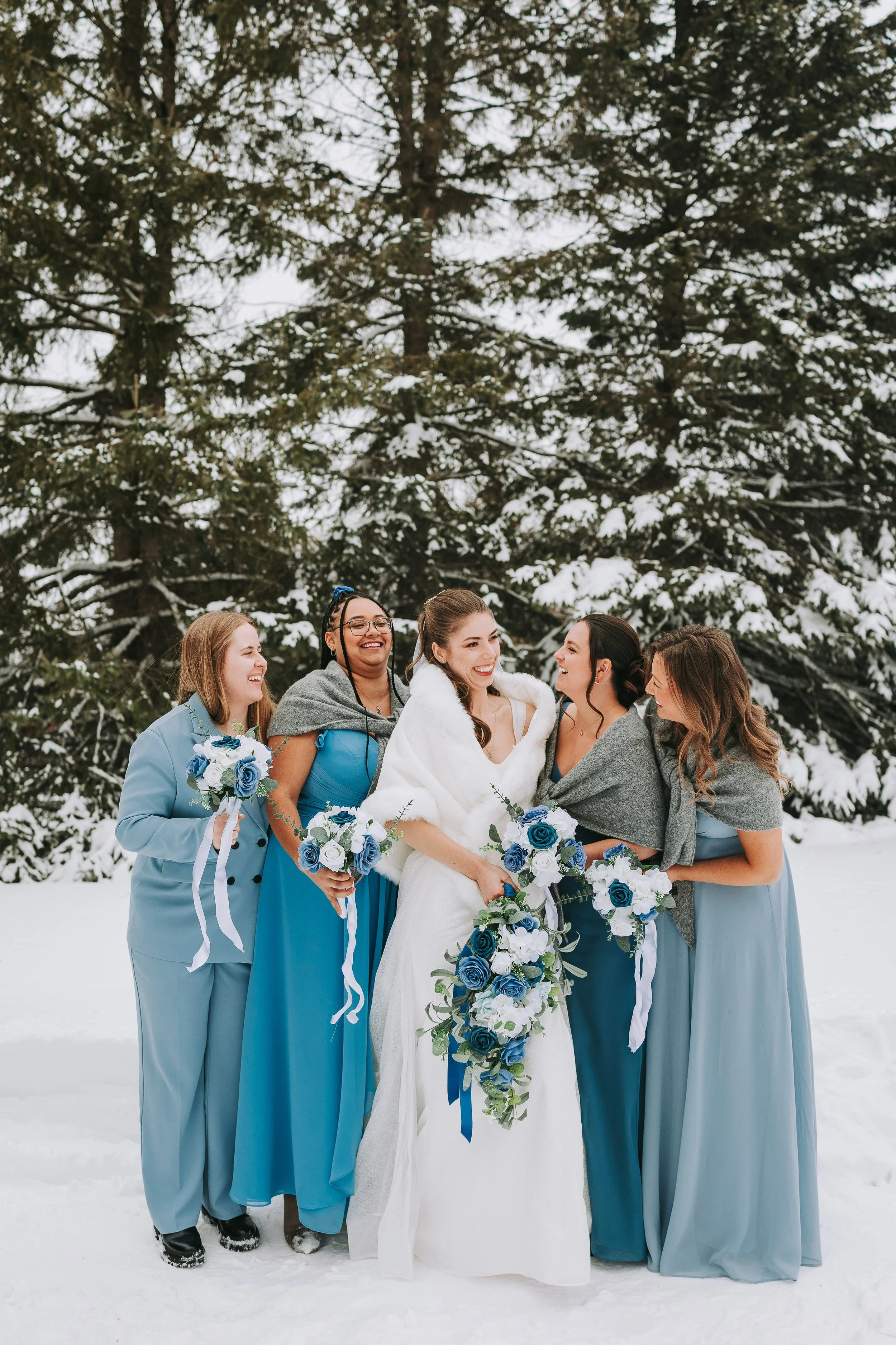 A bride in a white wedding dress and a white coat holding a cascading bouquet of blue and white roses, standing in snow with five women in blue dresses and gray shawls and jackets, all holding matching bouquets, smiling and laughing in a winter outdo