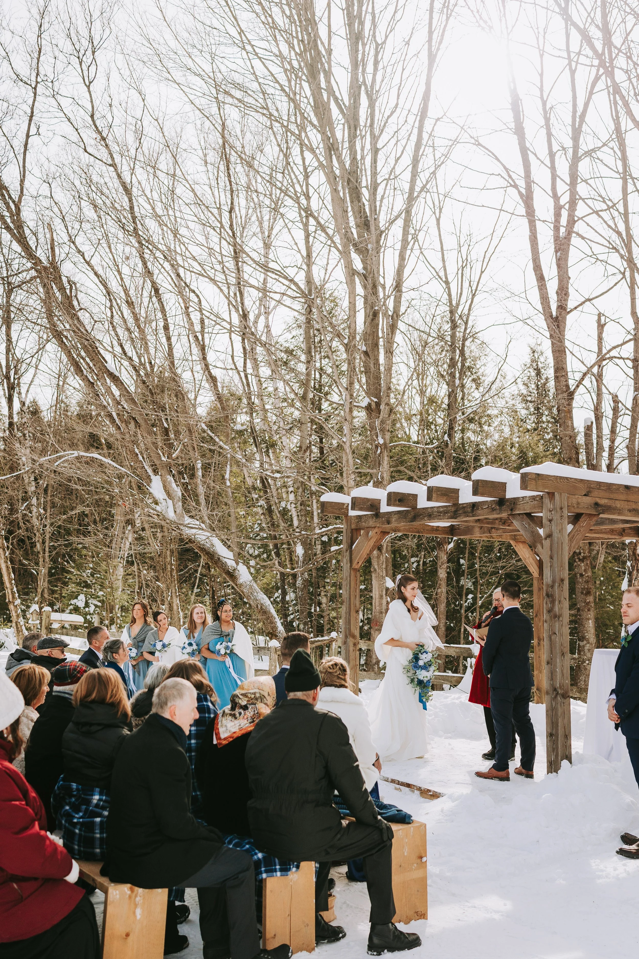 A winter outdoor wedding ceremony with a bride, groom, and officiant standing under a wooden arch, guests seated on wooden benches in snow, surrounded by bare trees.