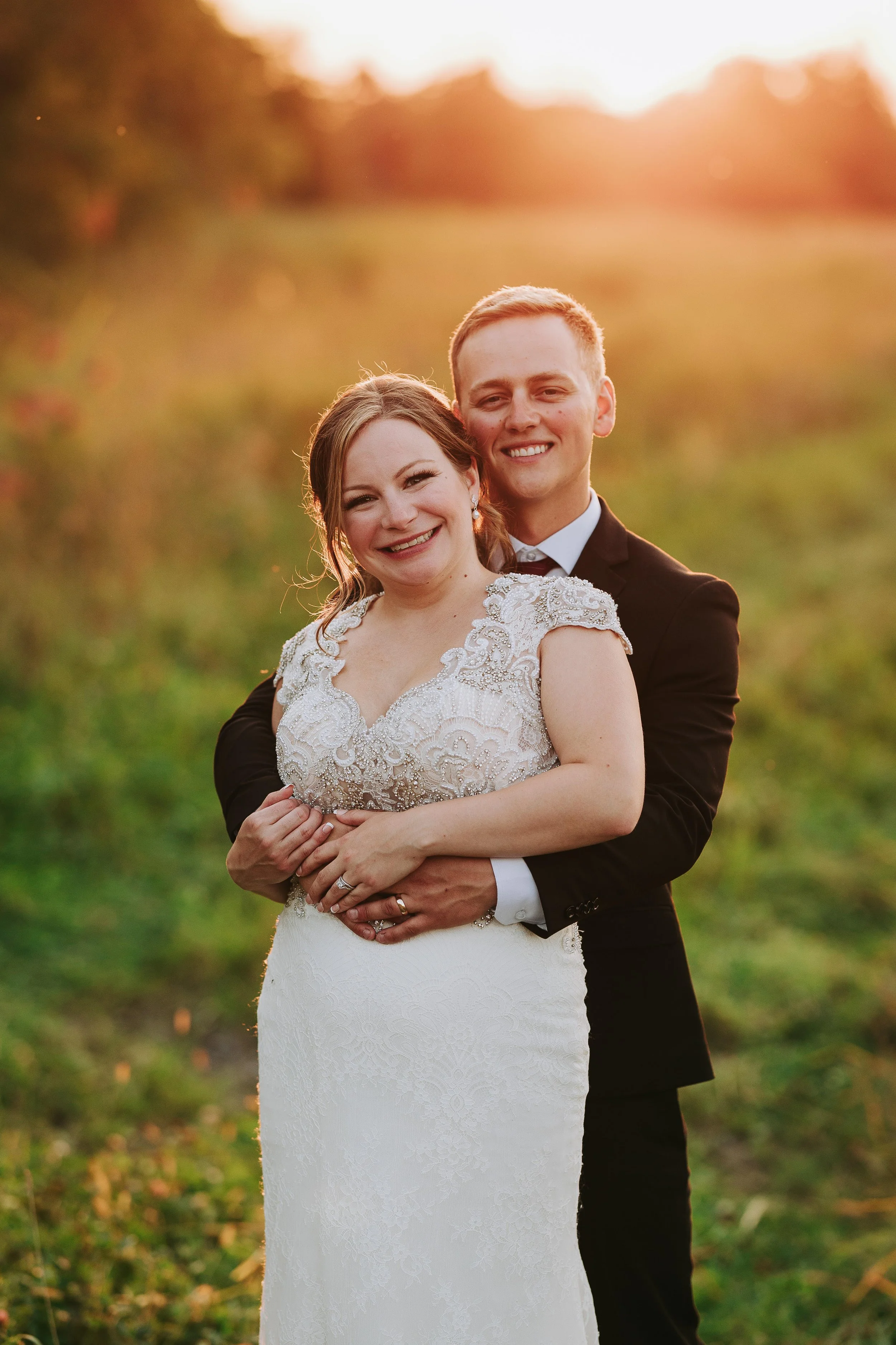 A newlywed couple hugging outdoors at sunset. The bride is wearing an elegant lace wedding gown, and the groom is in a black suit with a bow tie. They are smiling happily in a lush green area with soft sunlight behind them.